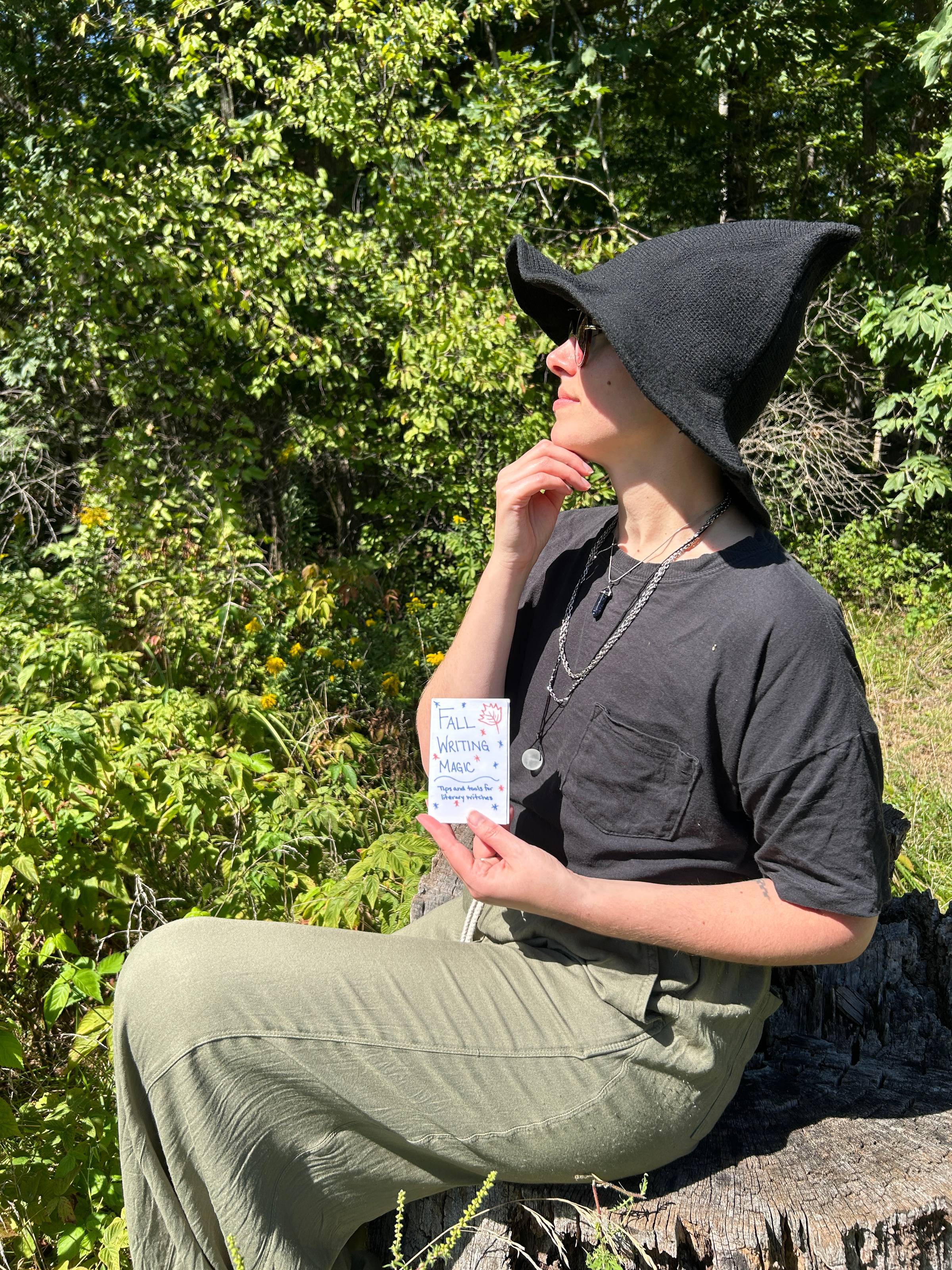 Bailey is wearing a black knit witch hat, black shirt, and green pants. They are sitting outside on a stump and holding up a copy of the zine, looking thoughtfully into the distance