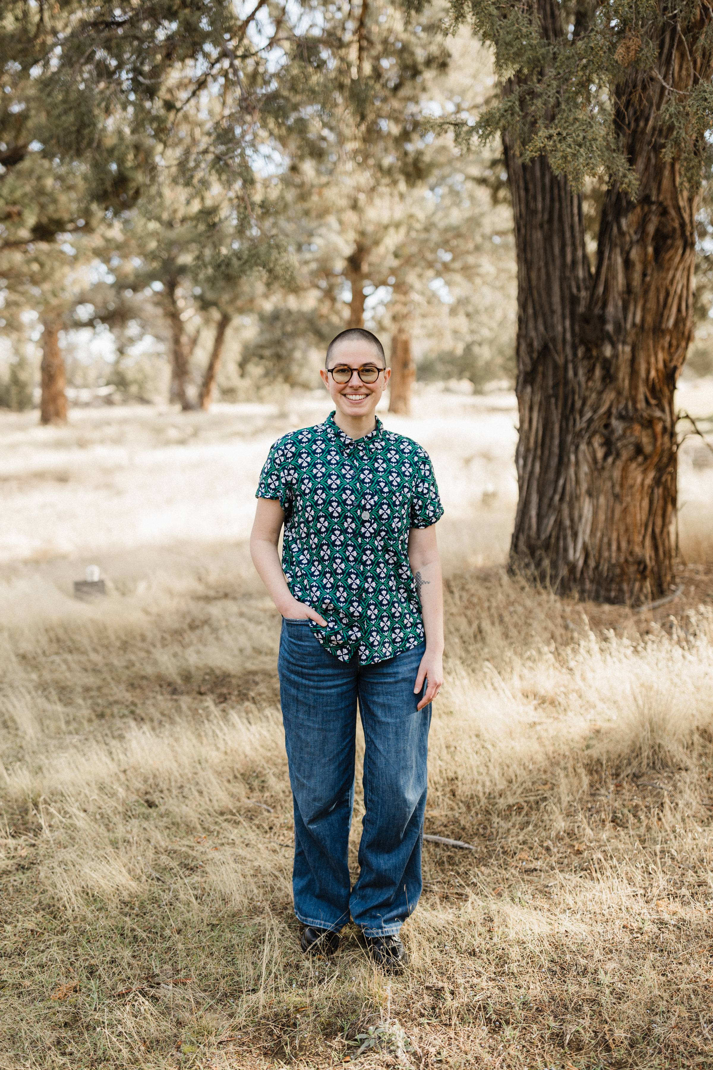 Bailey is a white nonbinary person with a shaved head. They are standing outside near a tree. They have glasses on and are wearing a green patterned shirt and jeans.