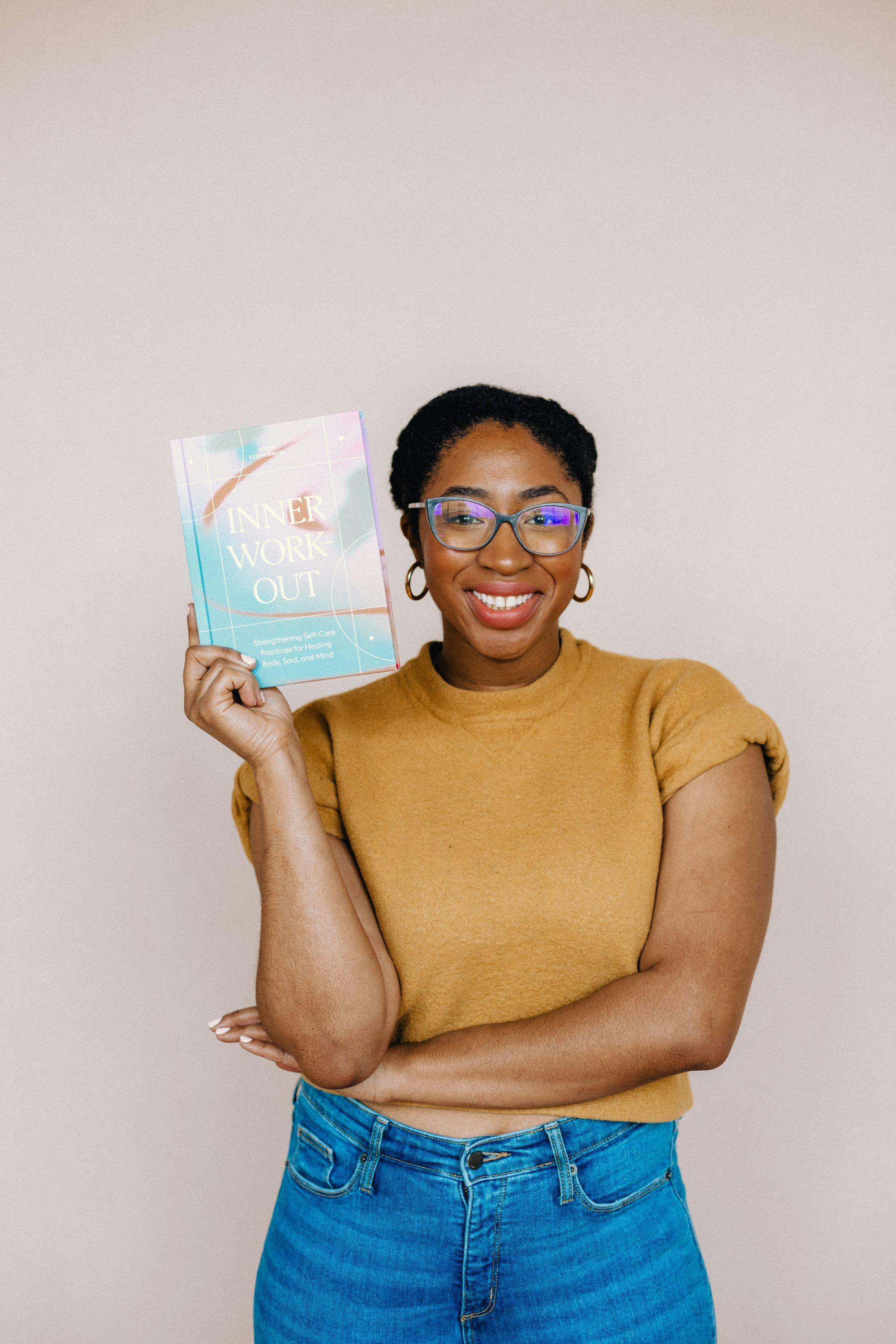 Taylor, a Black woman with glasses and wearing a yellow sweater, holds a copy of the book Inner Workout