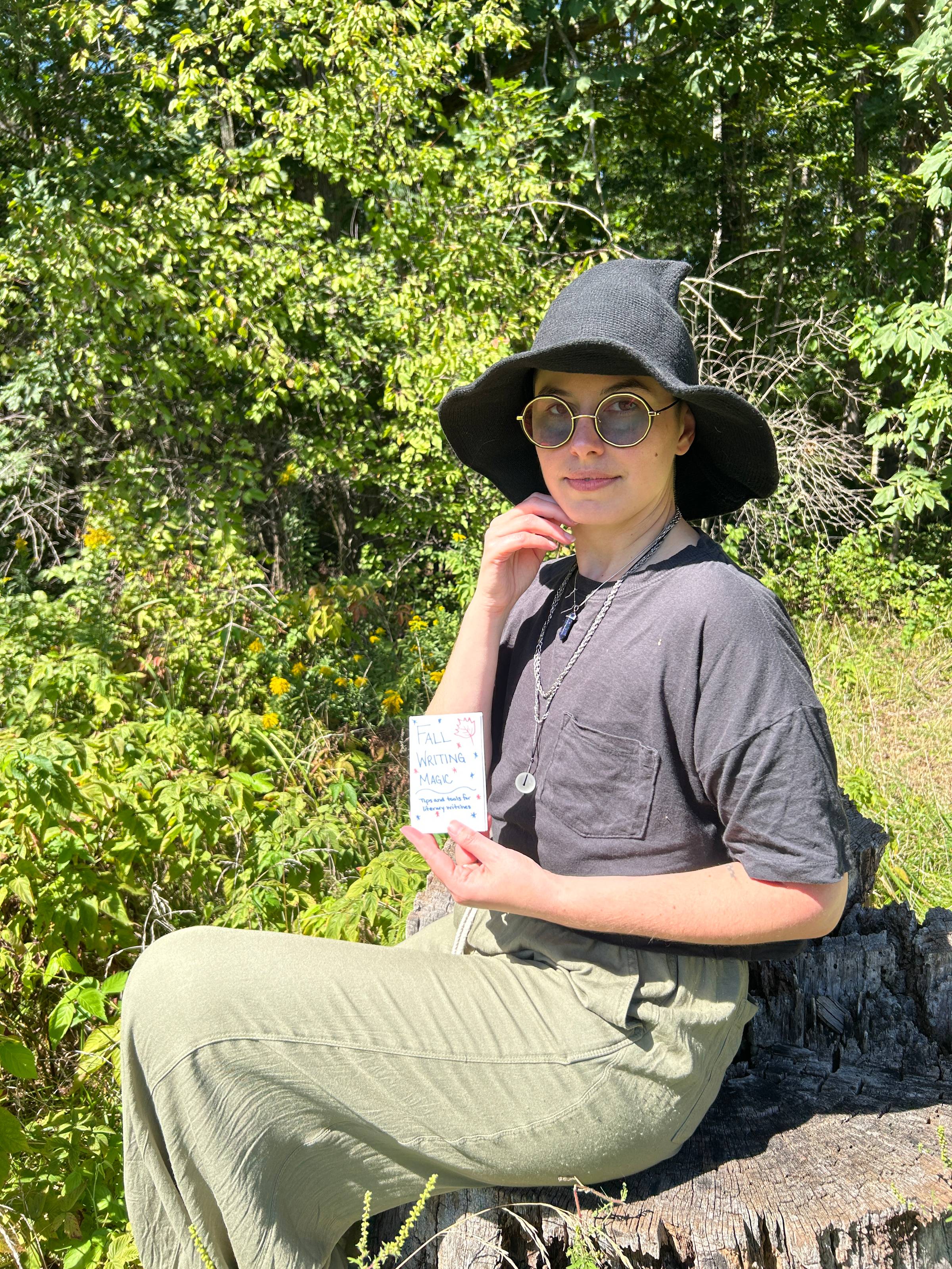 Bailey sits on a stump holding up a copy of the Fall Writing Magic zine. They are looking at the camera wearing a playful expression and a witch hat.