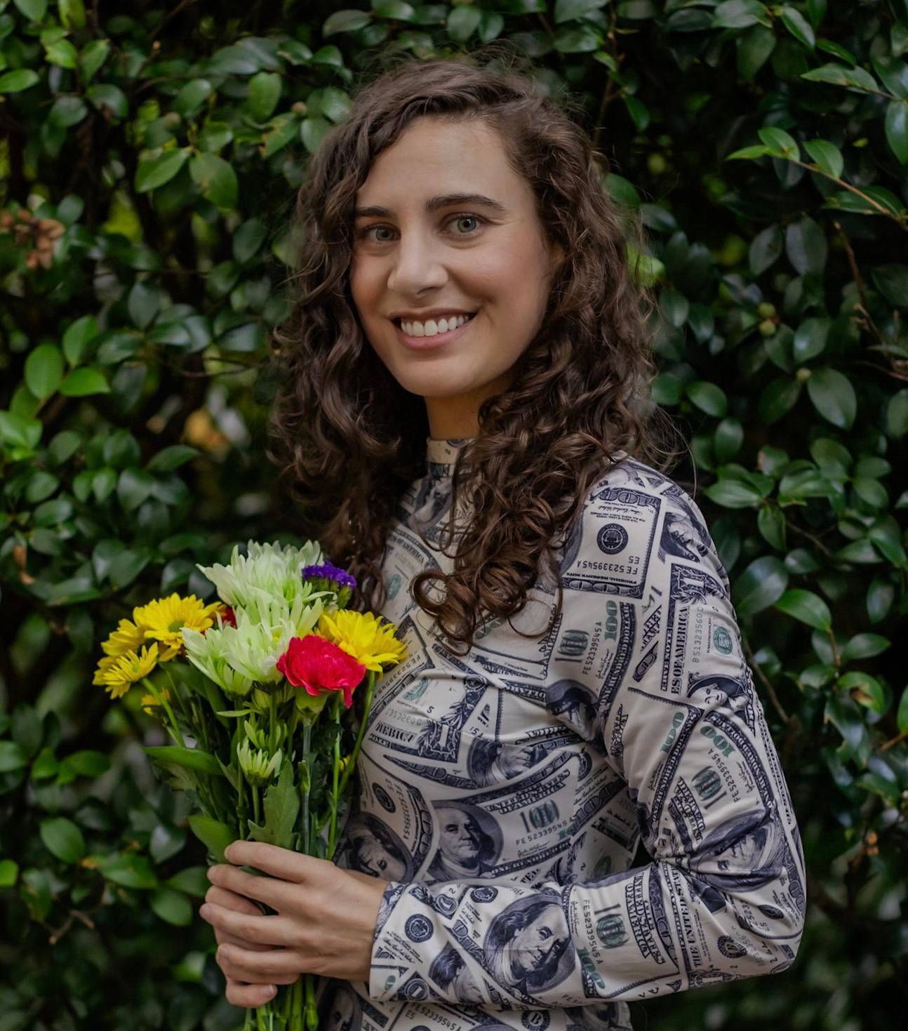 Kara Perez has curly brown hair. She is standing in front of a backdrop of greenery, holding a bouquet, and wearing a top printed with $100 bills