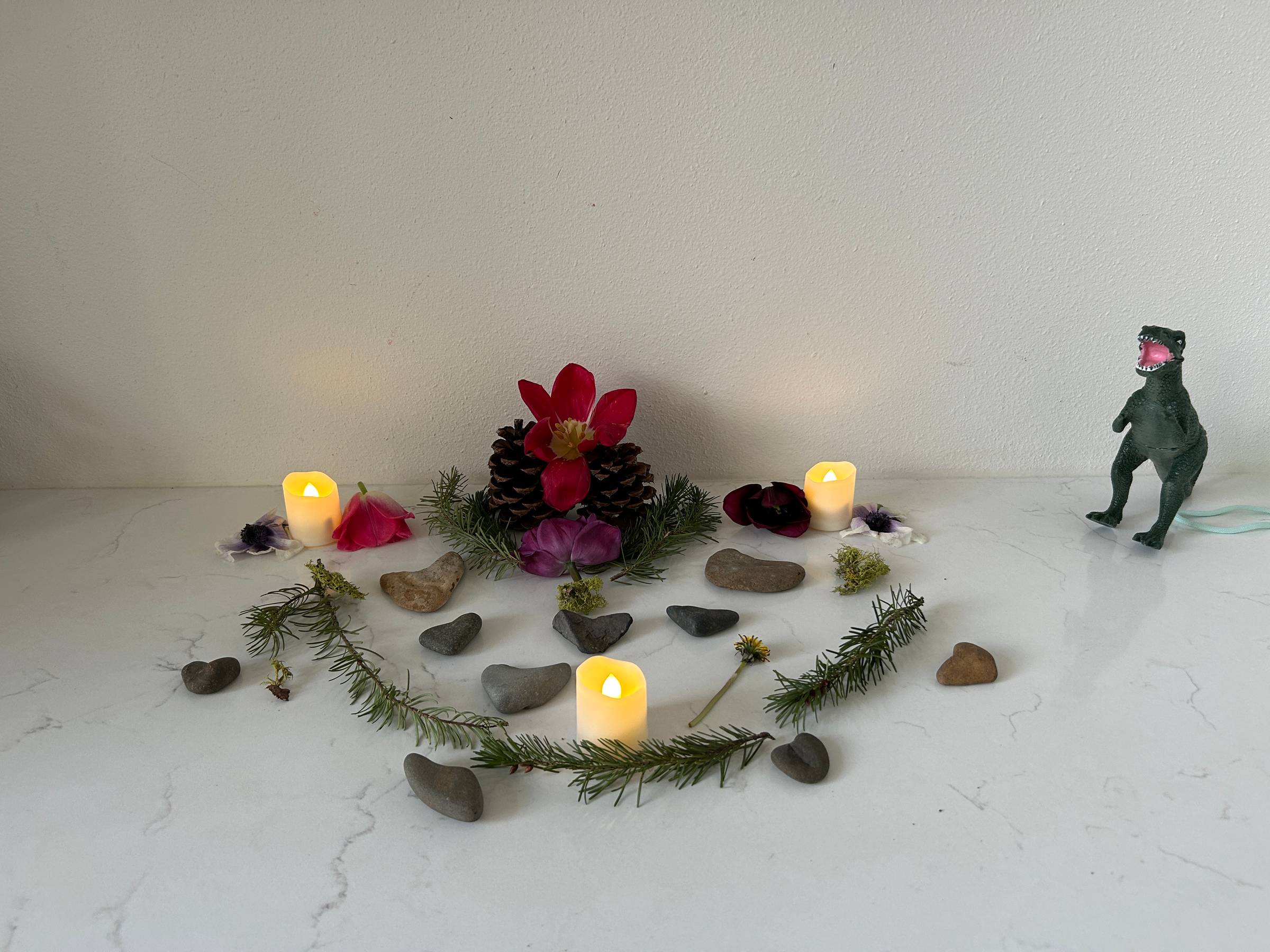 A collection of natural objects—pinecones, flowers, rocks, and evergreen branches—arranged in a symmetrical pattern with some battery-operated candles. A plastic dinosaur sits nearby.