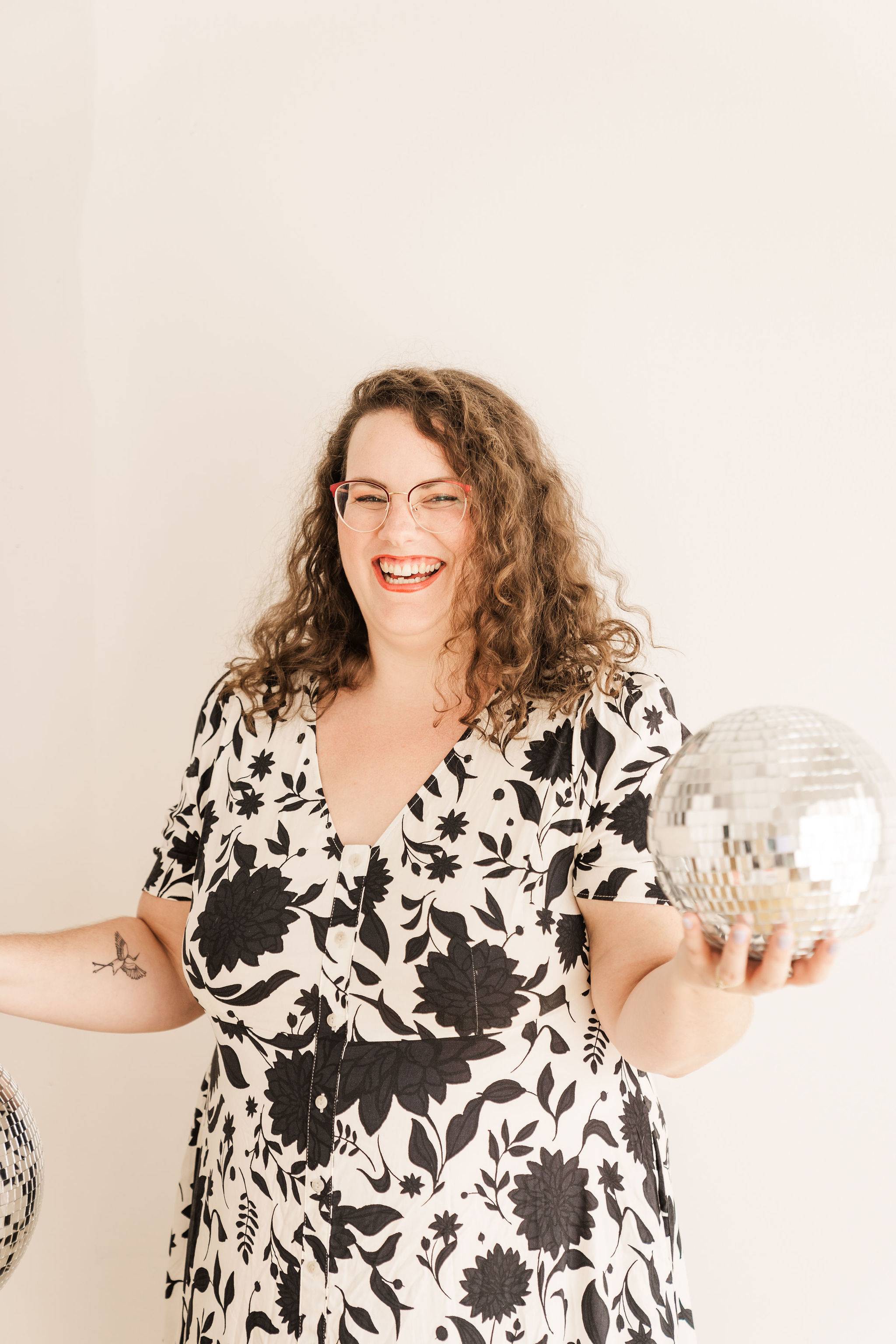 Amelia Hruby has wavy brown hair and is wearing a black and white printed dress. She is smiling widely and holding a disco ball in one hand.