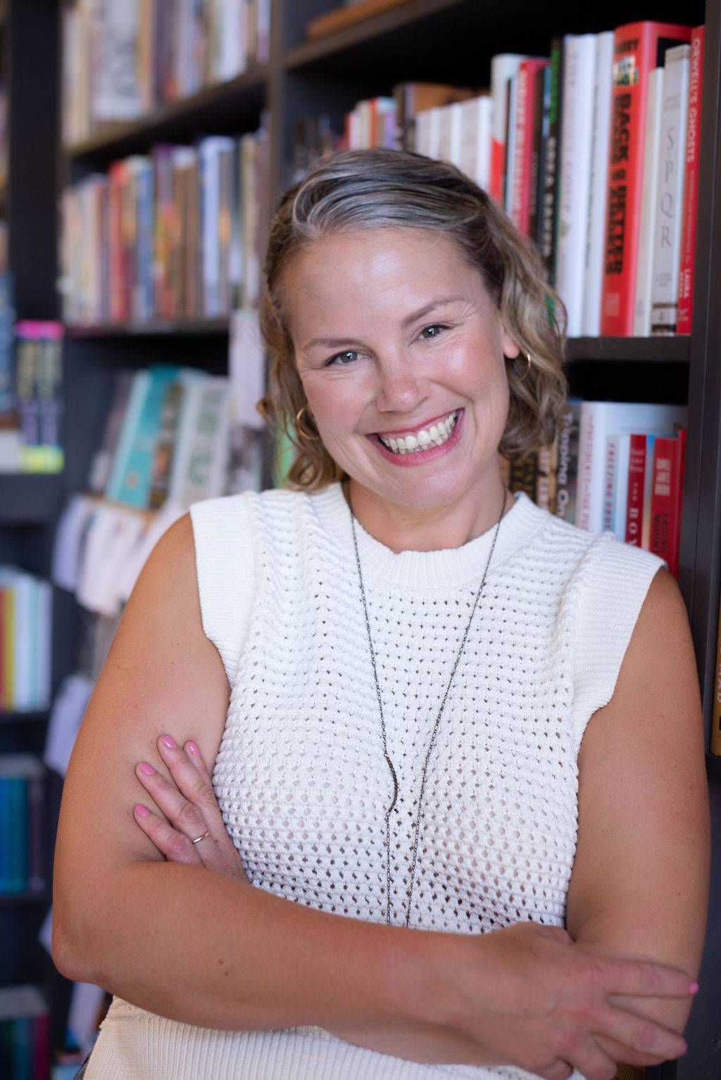 Meghan Stevenson is a white woman with blonde hair. She is wearing a knit white top and standing in front of a bookshelf.