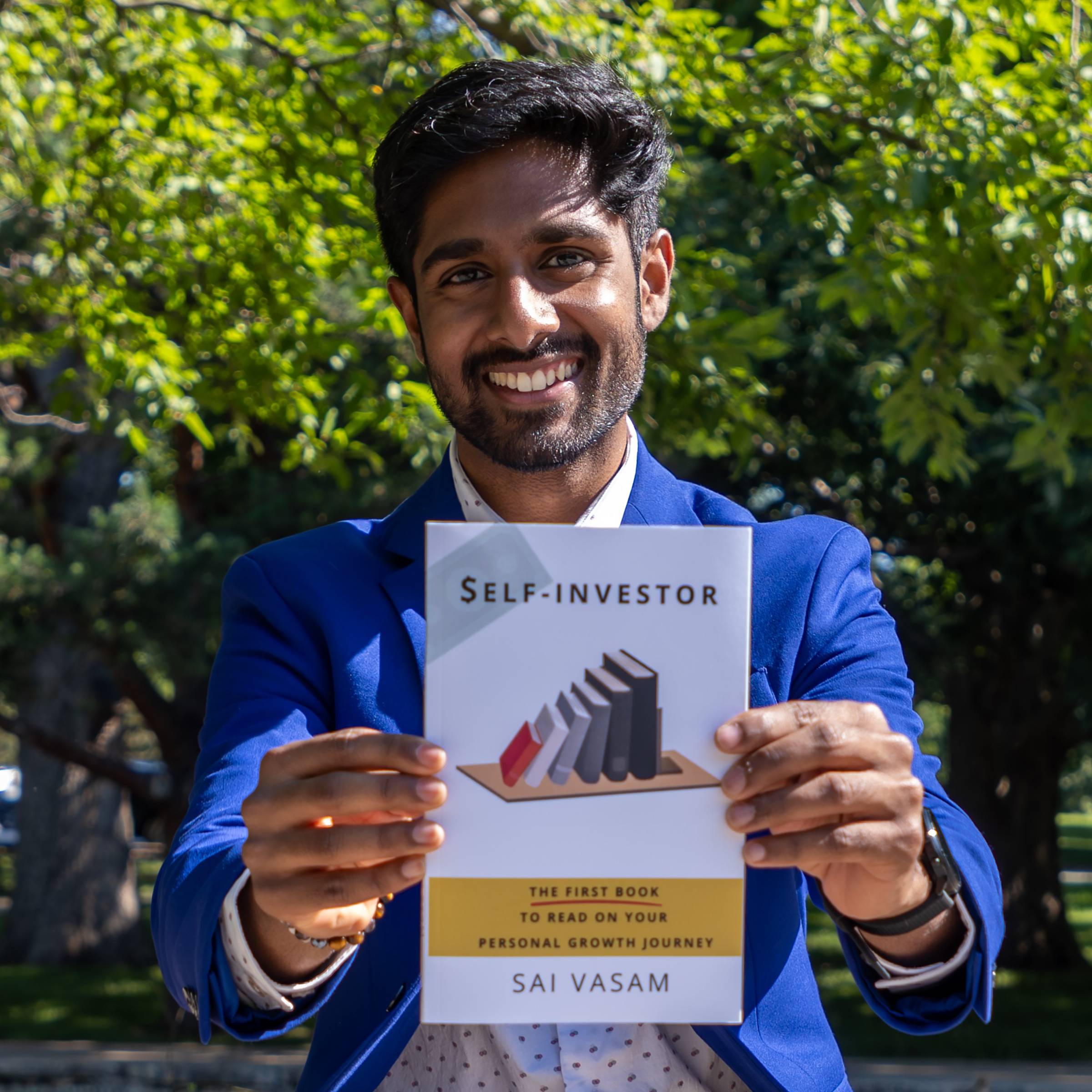 Sai is standing outside wearing a bright blue jacket over a polka dotted button-down and holding up a copy of his book.