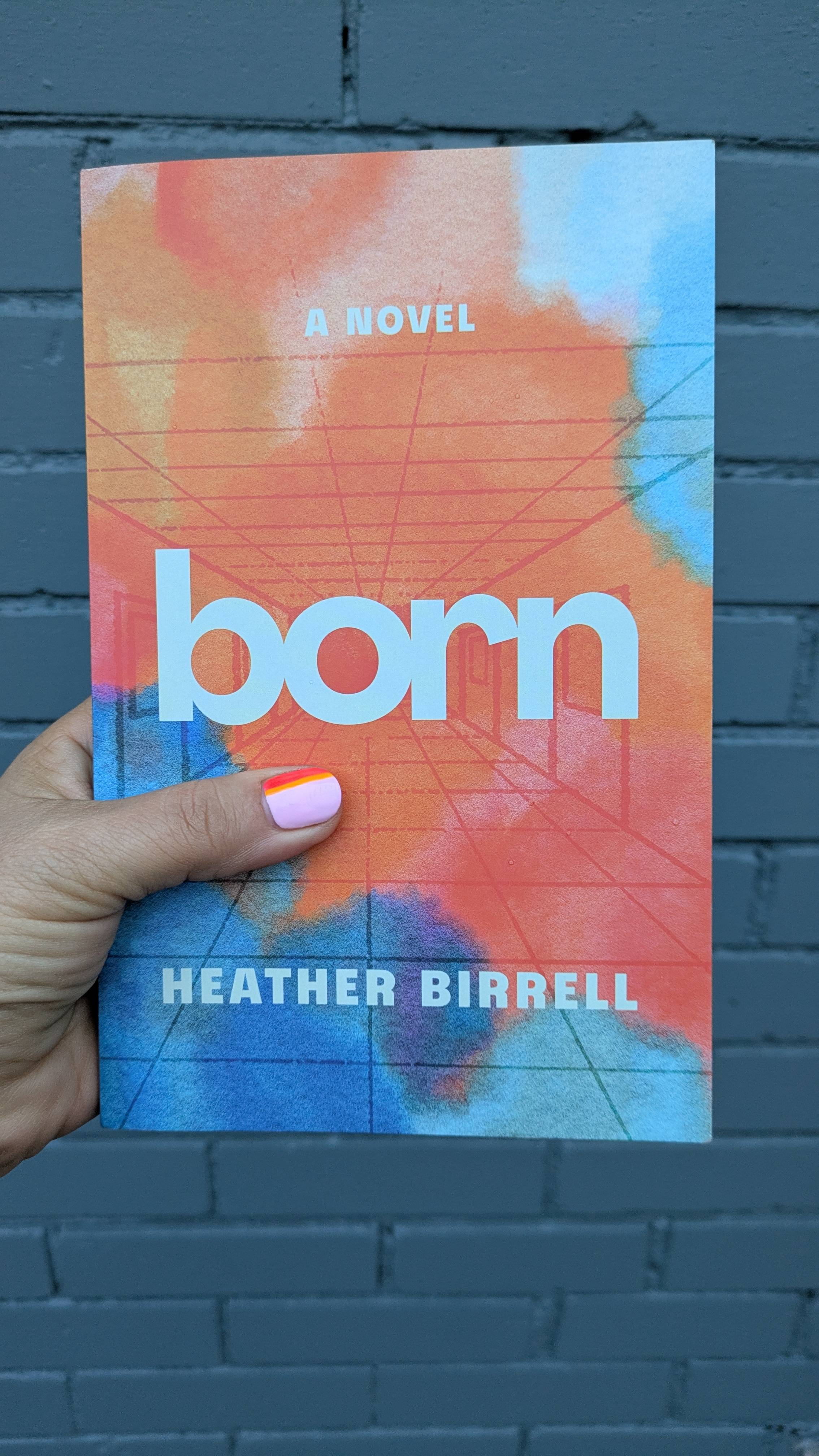 A picture of someone's hand holding a copy of Born in front of a blue-gray brick wall. The person's thumbnail is painted to match the abstract colors on the cover. The cover image features a drawing of a school hallway and the book's title and Heather's name.