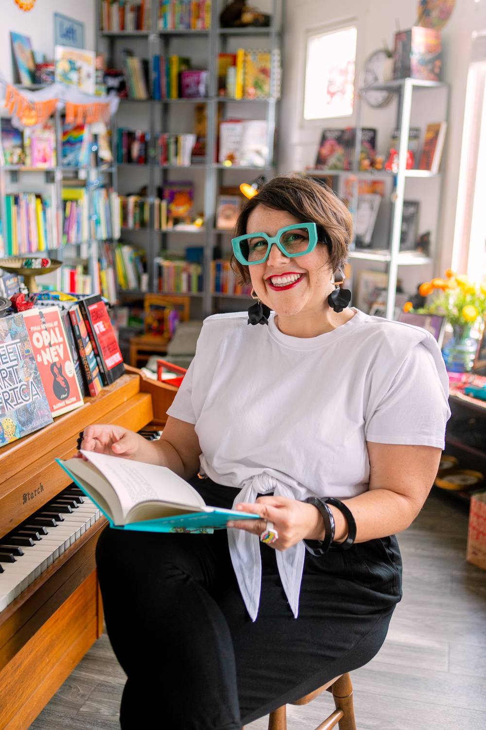 Lauren Marie Fleming is a white woman with short brown hair. She is wearing large, bright turquoise glasses and sitting with a book in her lap next to a piano.