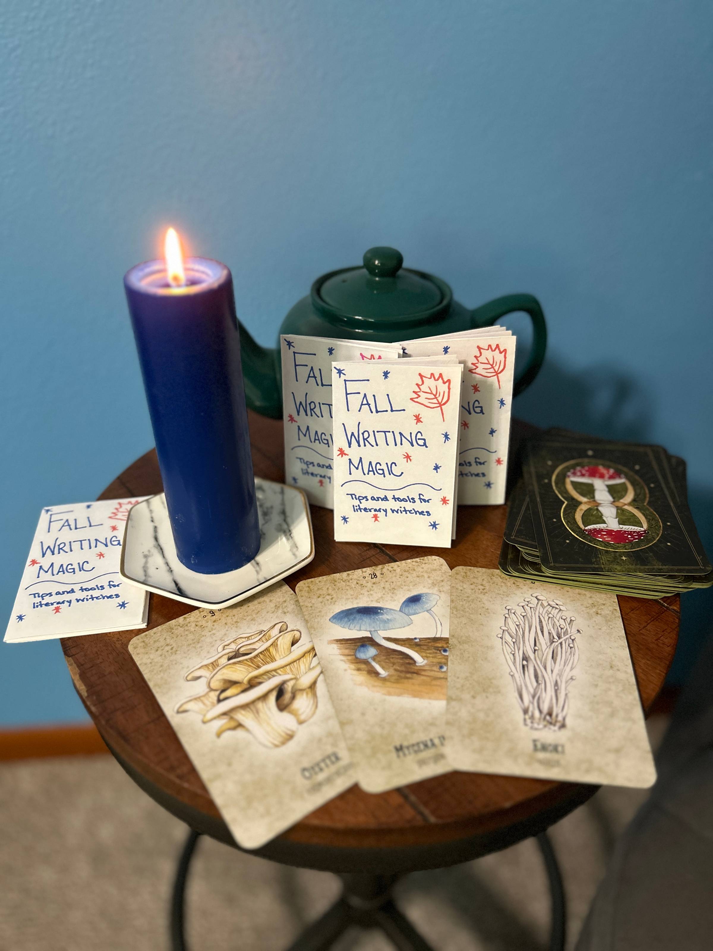 The Fall Writing Magic zine sits on a table with a spread of oracle cards, a lit candle, and a teapot