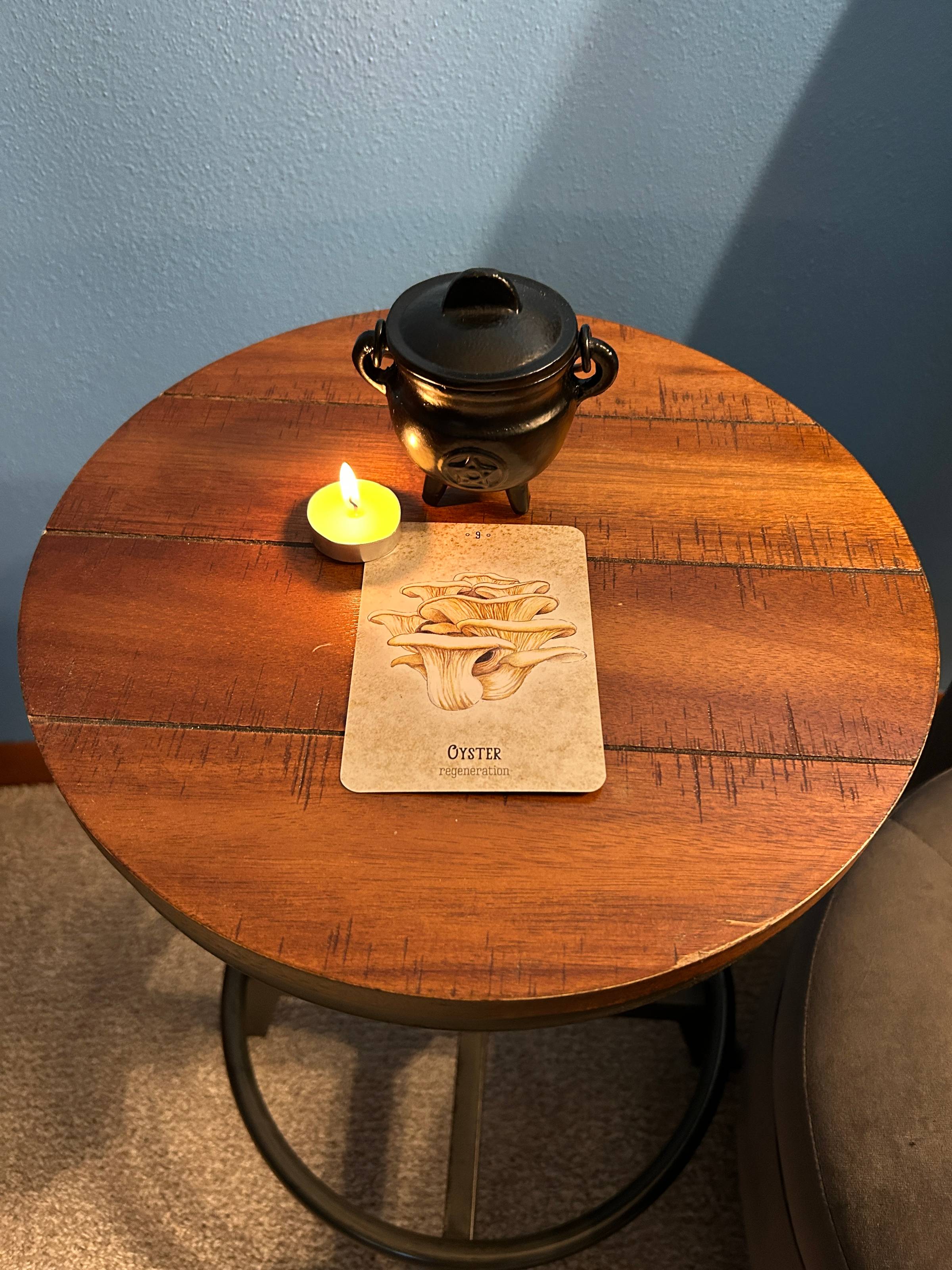 An illustratd oyster mushroom oracle card sits on a small round stool next to a tealight candle and a small cast-iron cauldron.