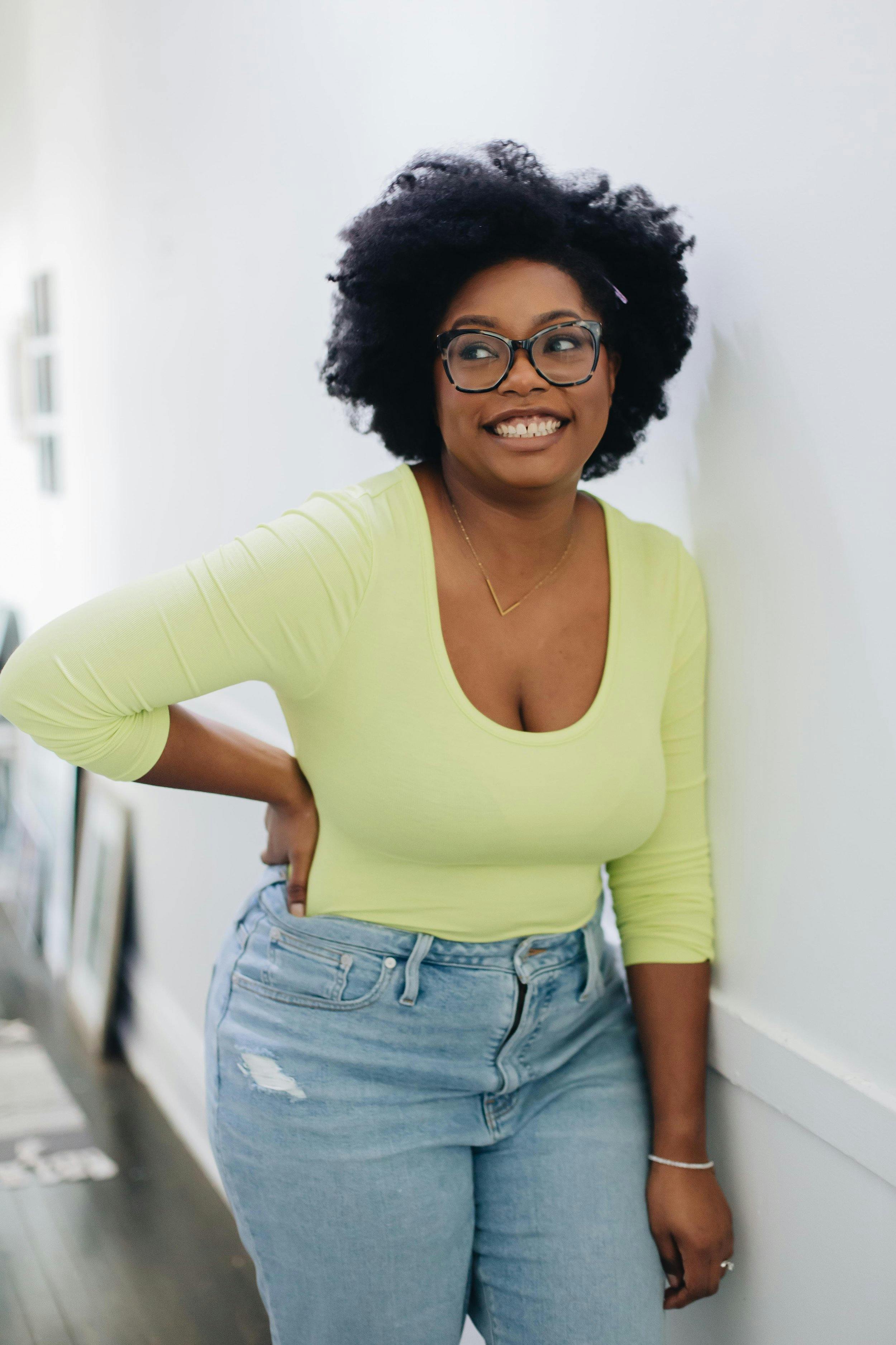 L'Oreal is a Black woman wearing a light green shirt and jeans. She is smiling as she leans against a wall. 