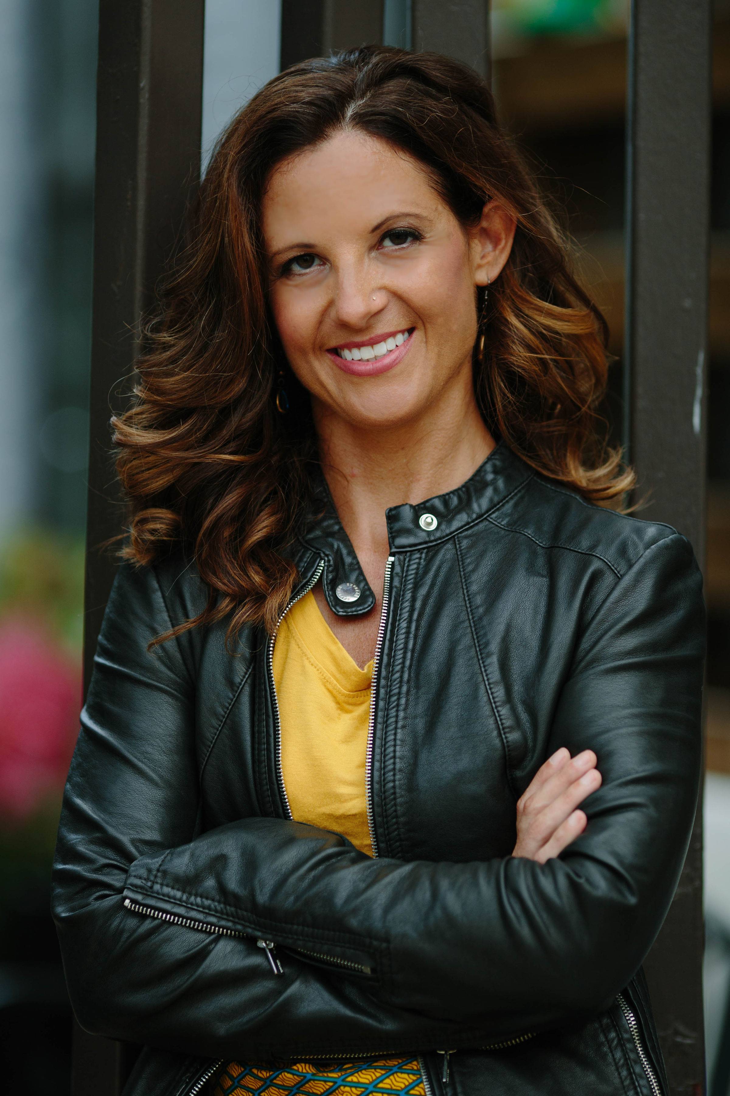 Emily Crookston is wearing a dark leather jacket over a bright yellow shirt. She is standing against a glass backdrop with her arms crossed and smiling.