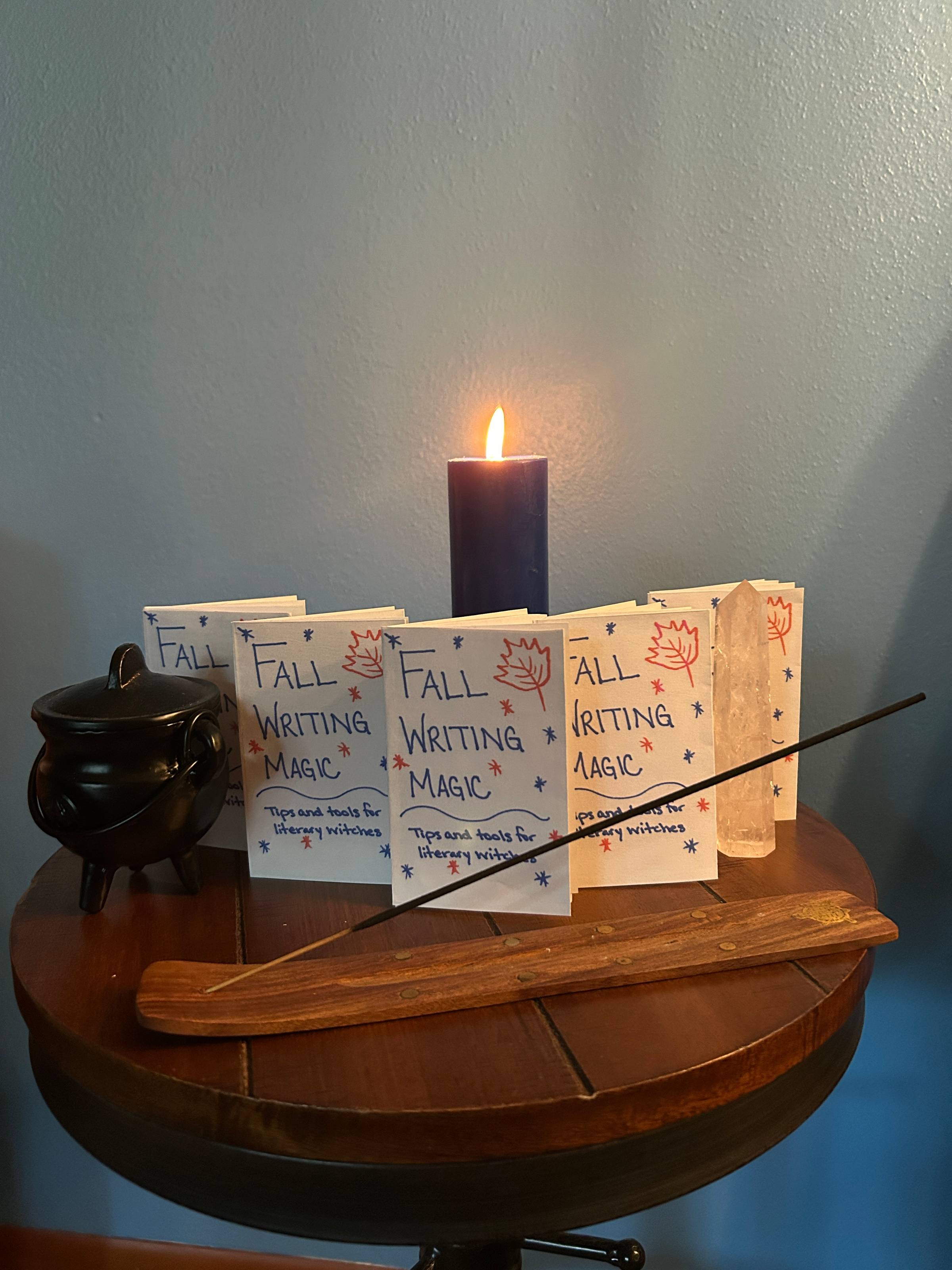 5 copies of the Fall Writing Magic zine sit on a table with a small iron cauldron, a lit candle, and a stick of incense