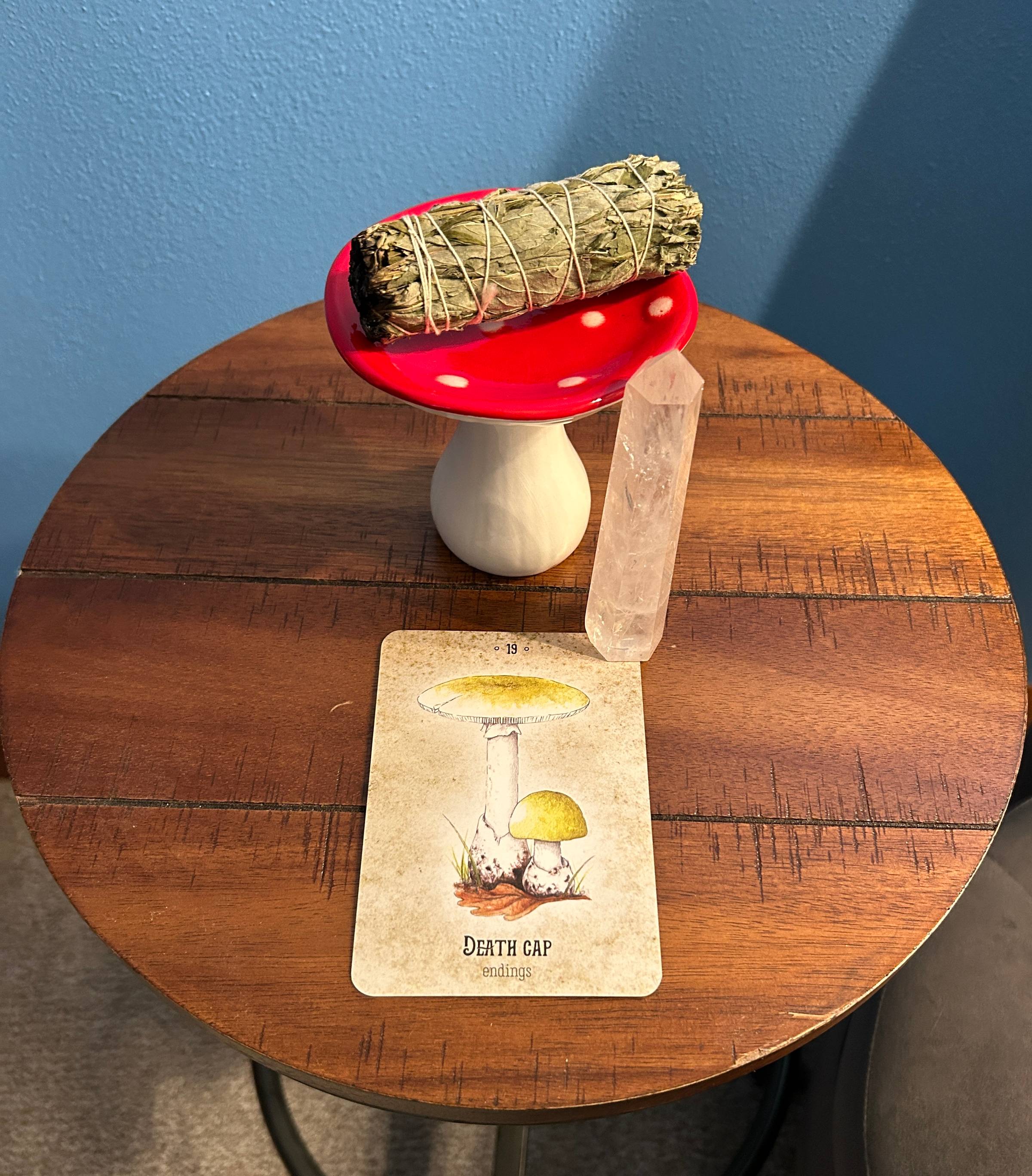 A death cap oracle card sits on a small round table. The same white crystal from the previous image is on the card. A white and red mushroom stand holds a bundle of mugwort behind the card.