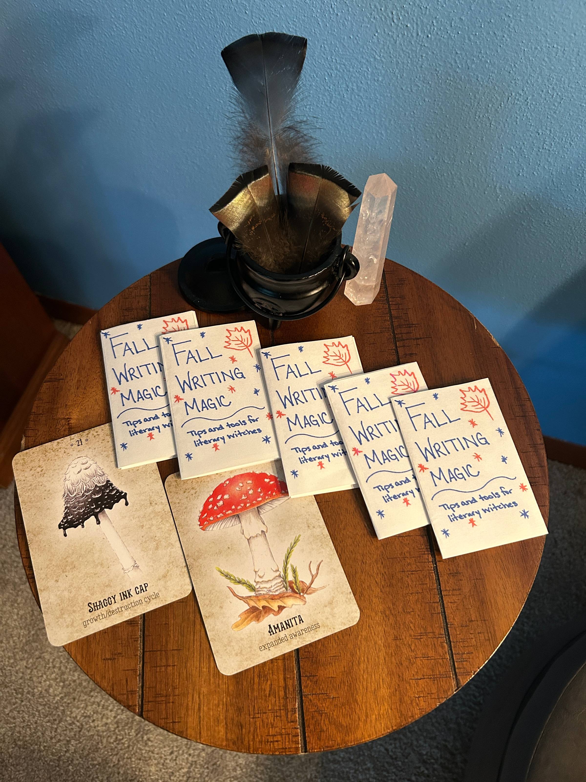 5 copies of the Fall Writing Magic zine sit on a round table next to two oracle cards. Behind them is a crystal and a collection of feathers in a small metal cauldron.