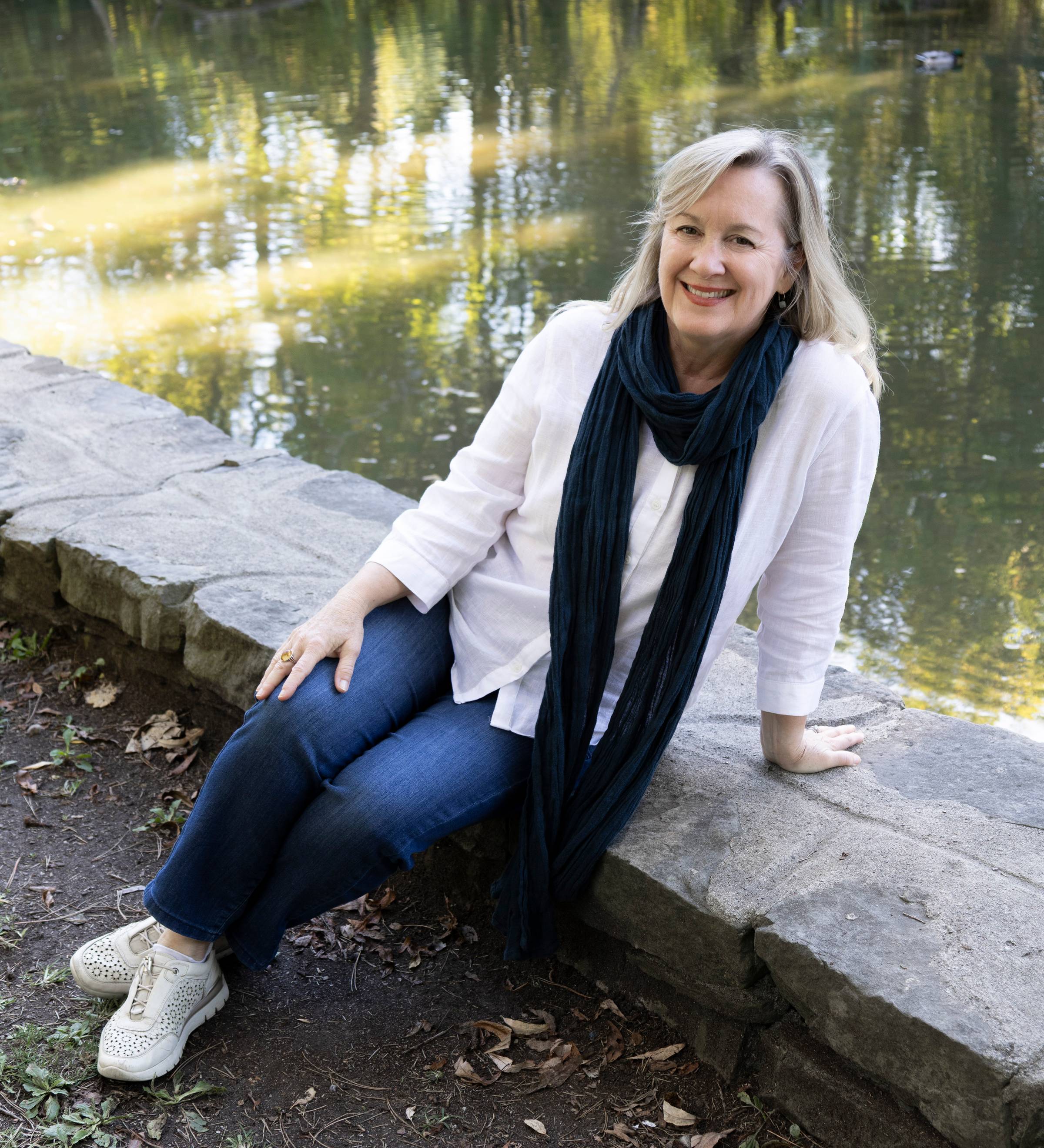 Christine is wearing a long black scarf, white top, and jeans. She is sitting at the edge of a body of water.
