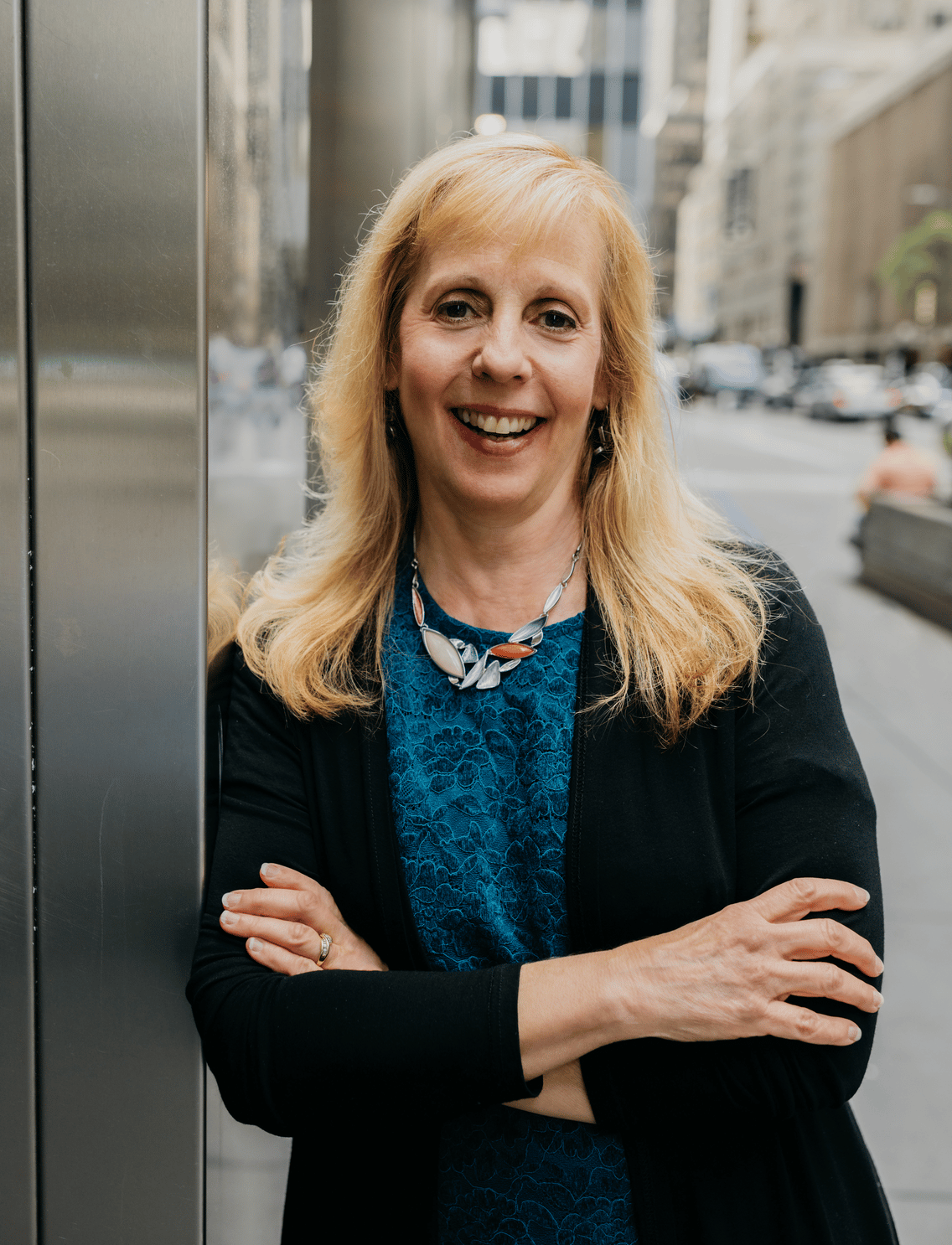 Meryl is a white woman with long blonde hair. She is wearing a dark blue shirt and a black blazer and leaning against a building near a city street.