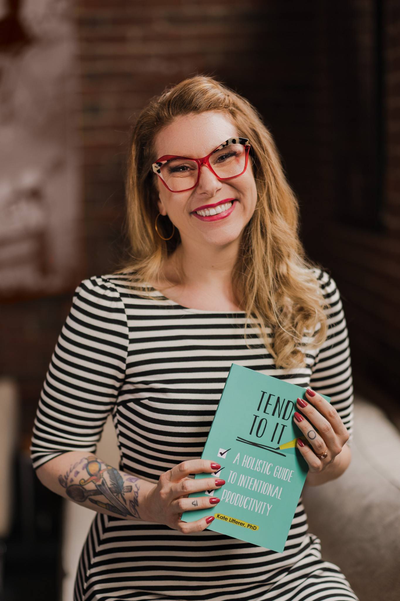 Dr. Kate Henry has long, curly blonde hair and is wearing a pair of glasses with bright red rims. She is holding a copy of her book and smiling.