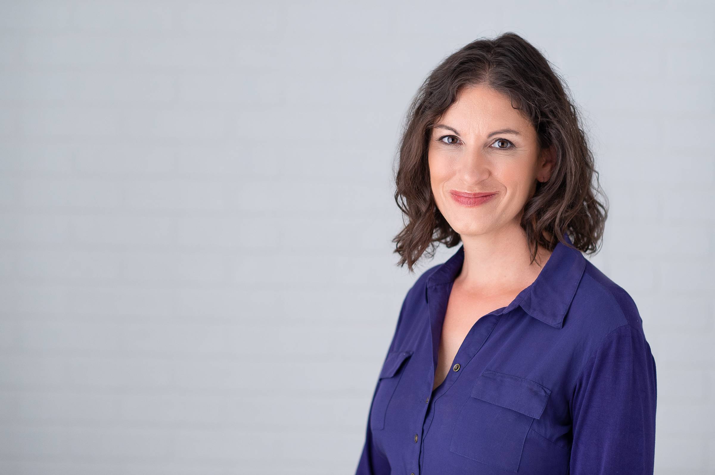 Heidi has shoulder-length wavy brown hair. She is wearing a dark blue top and standing in front of a white brick wall.