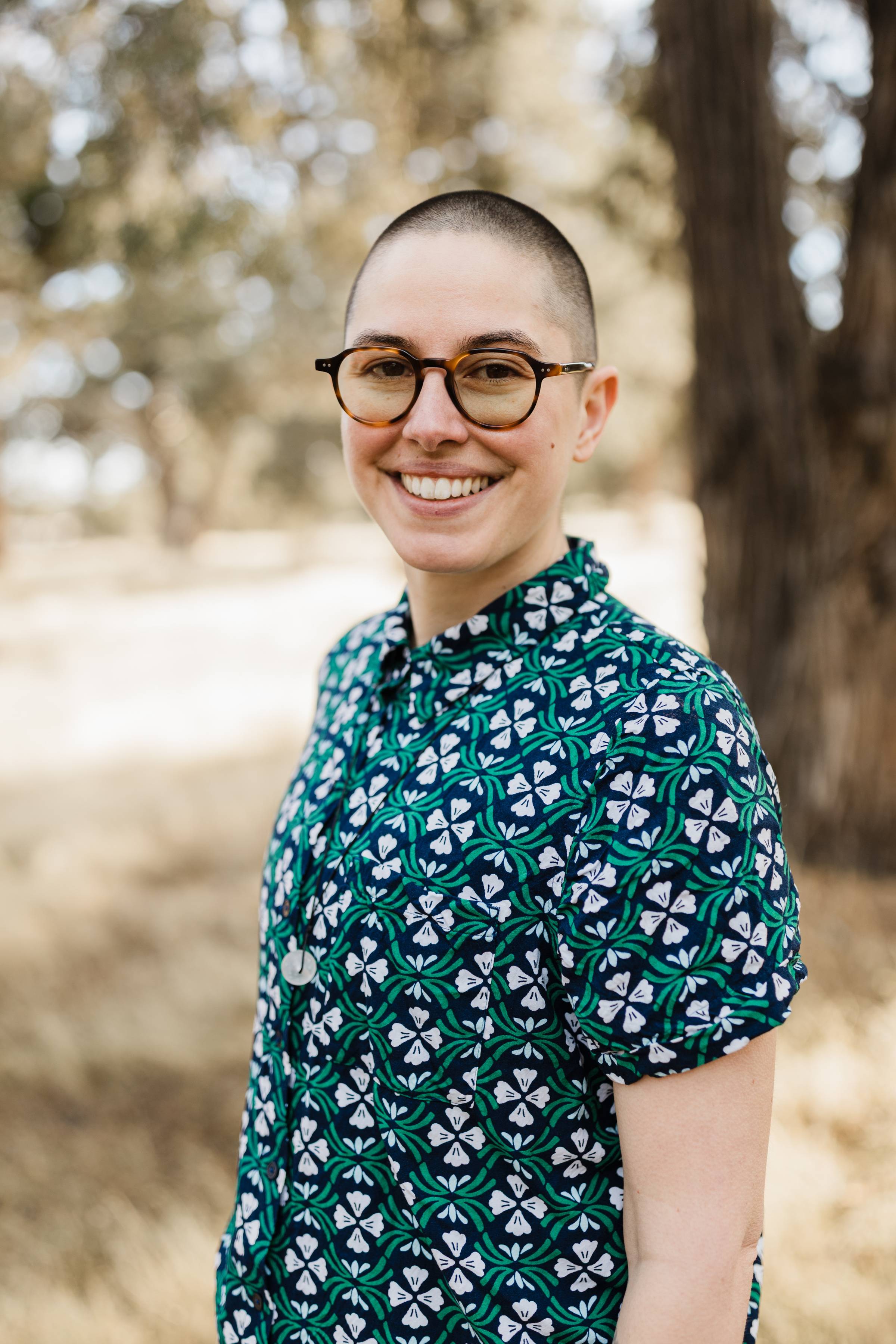 Bailey is a white person wearing a short-sleeved collared shirt. Their head is shaved and they wear glasses.