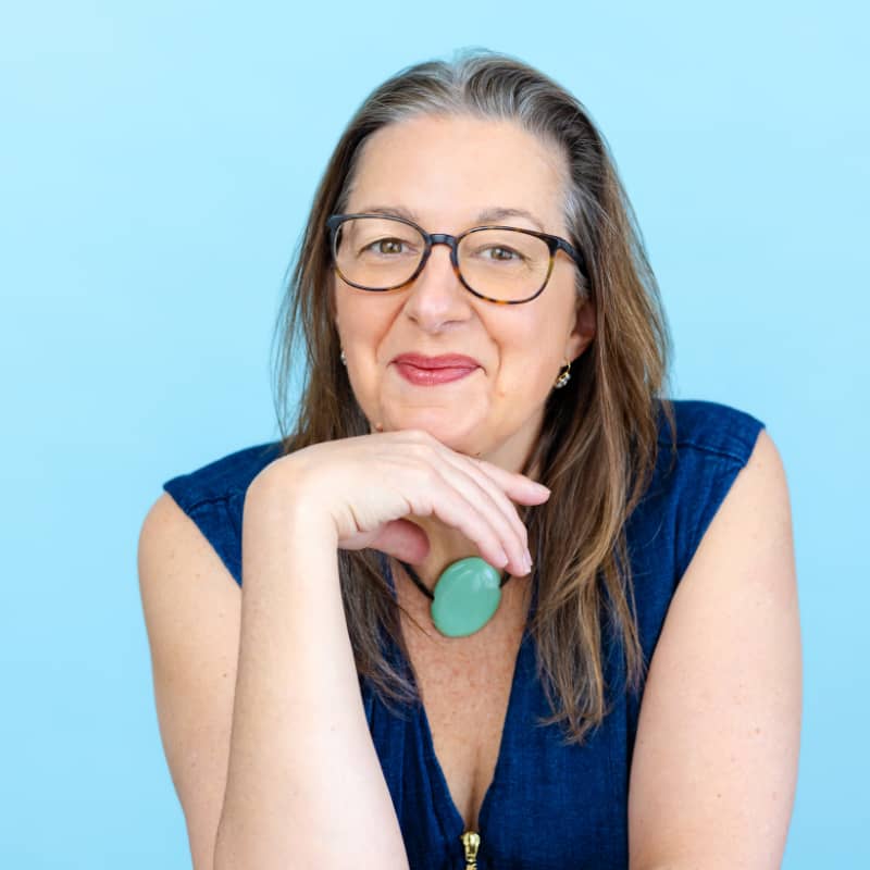 Kristen is sitting in front of a light blue background. She has her chin propped on her hand and is smiling at the camera.