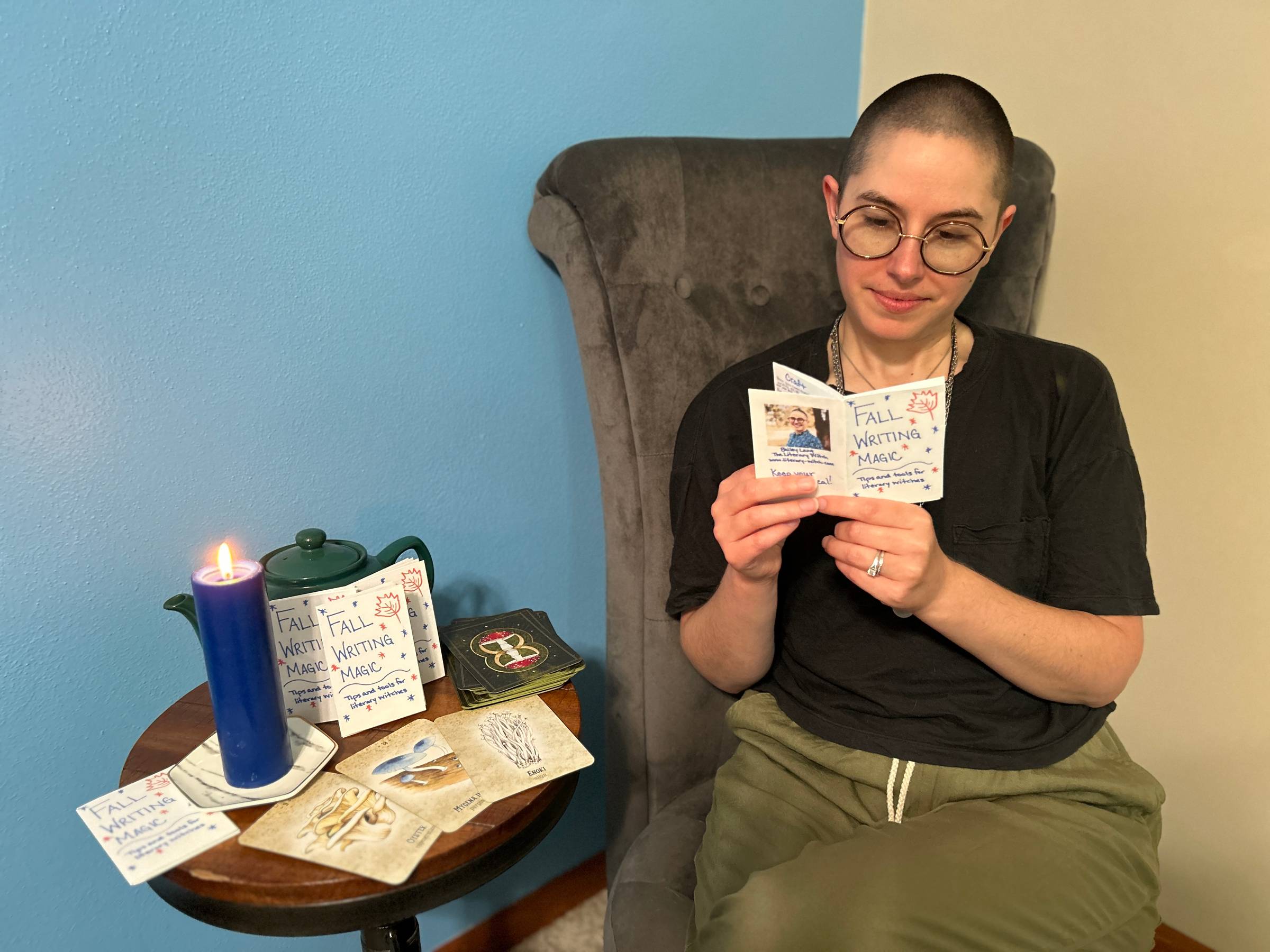Bailey is sitting in a gray chair reading a copy of the Fall Writing Magic zine. More copies of the zine sit on a small table next to them with a lit candle, some oracle cards, and a teapot.