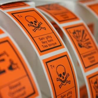 Rolls of bright orange warning stickers. Most are blurred. The one in focus has a skull and crossbones and the text "Very toxic" in three different languages.