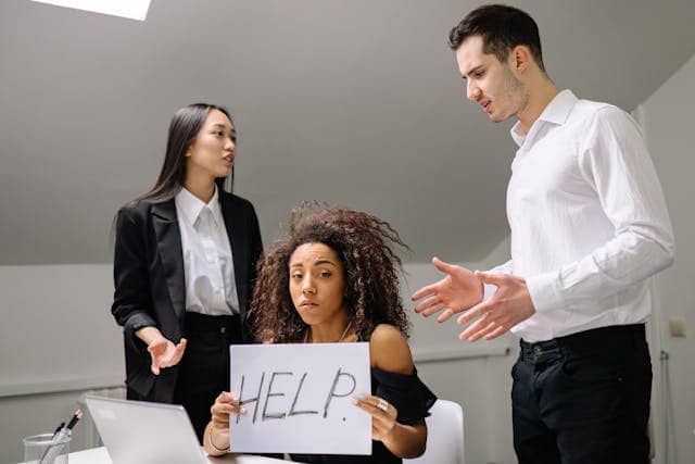 Three office workers. One is sitting, looking at the camera, and holding a sign that says "help". The two others are standing in positions that suggest a tense situation.