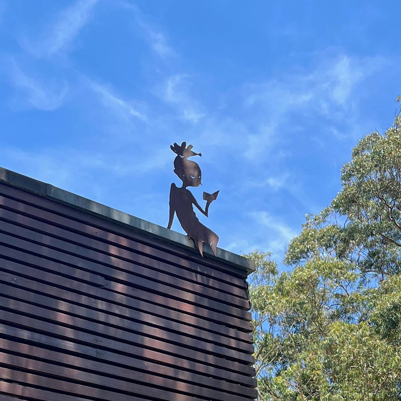 Photo of a metal cutout sculpture of a person sitting on the edge of a roof, reading a book. Background is blue sky and a green tree top.
