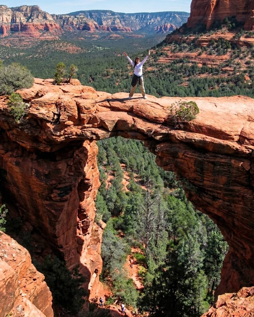 Jackie standing on the natural sandstone arch of Devil's Bridge with arms raised, dramatic canyon vista and red rock formations in the background
