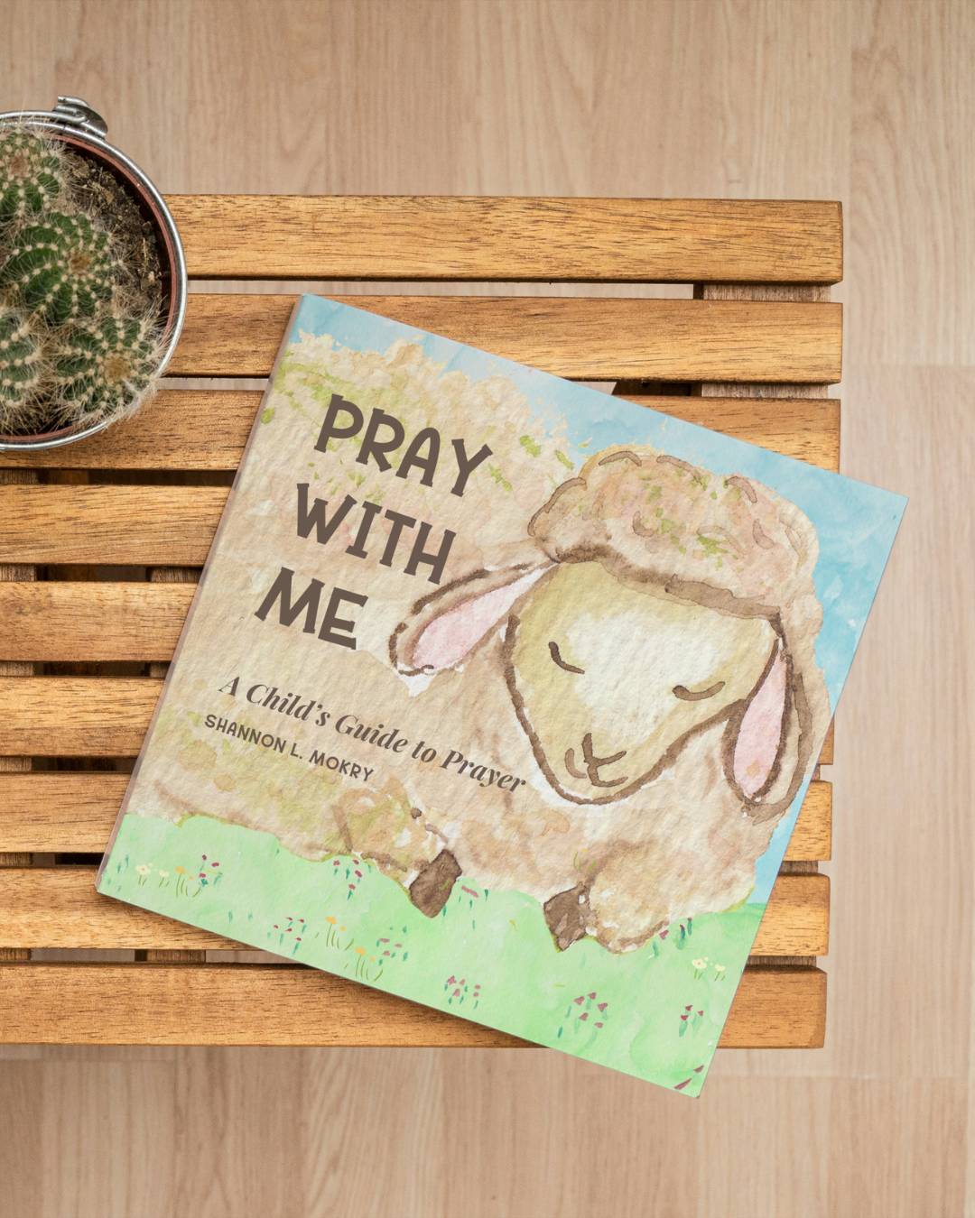 A wooden tables holds a book. The cover pictures a beige lamb on green grass and the words "Pray with Me" in large font under it states " A child's Guide to Prayer." by Shannon L. Mokry