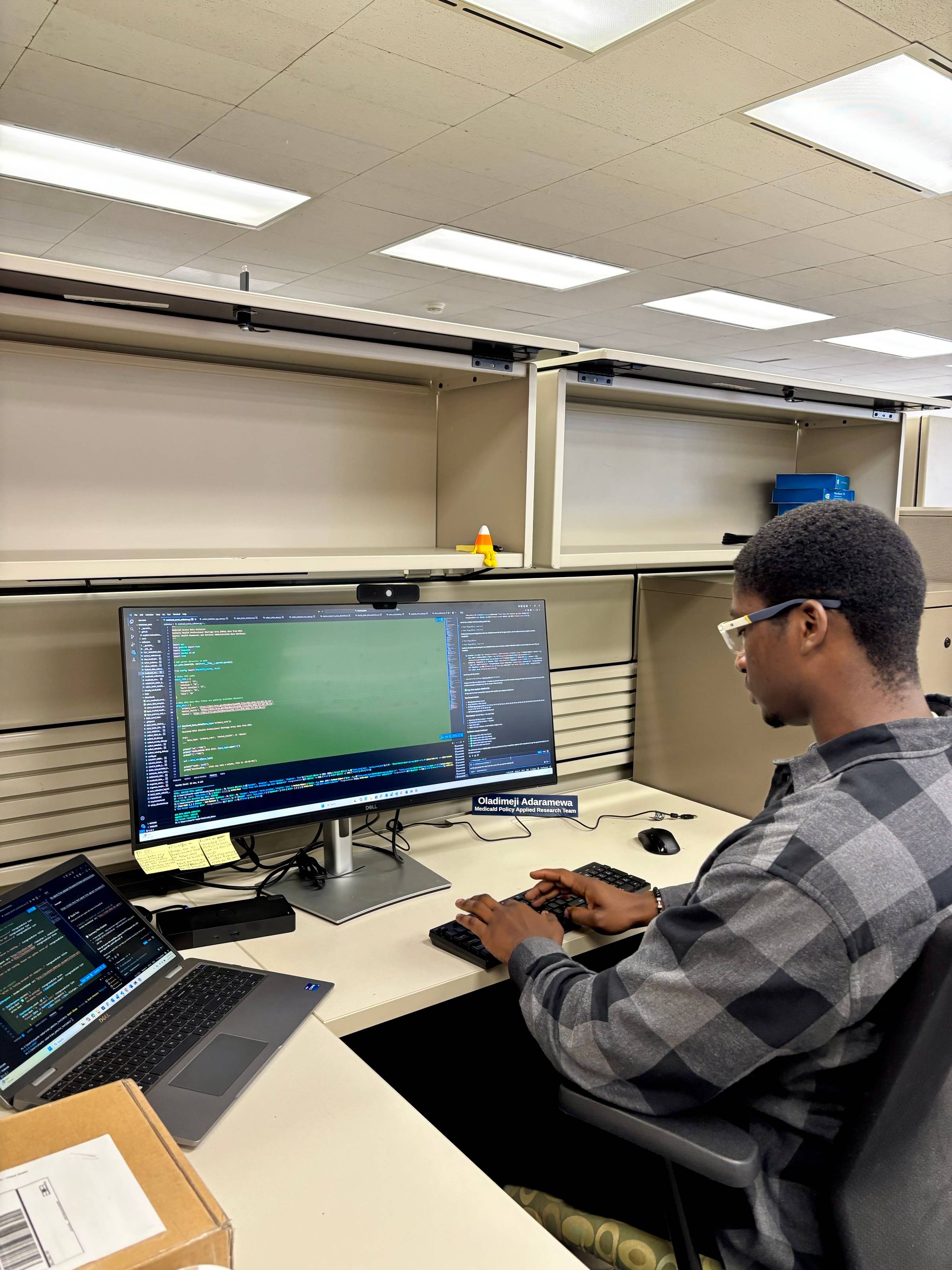 Dimeji Adaramewa working at his desk at the MPART office, with code displayed on a large monitor and his nameplate reading Medicaid Policy Applied Research Team