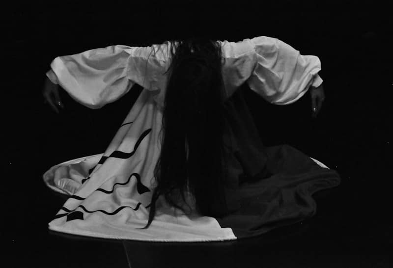 A person with long dark hair bowing in a white dress with black patterns