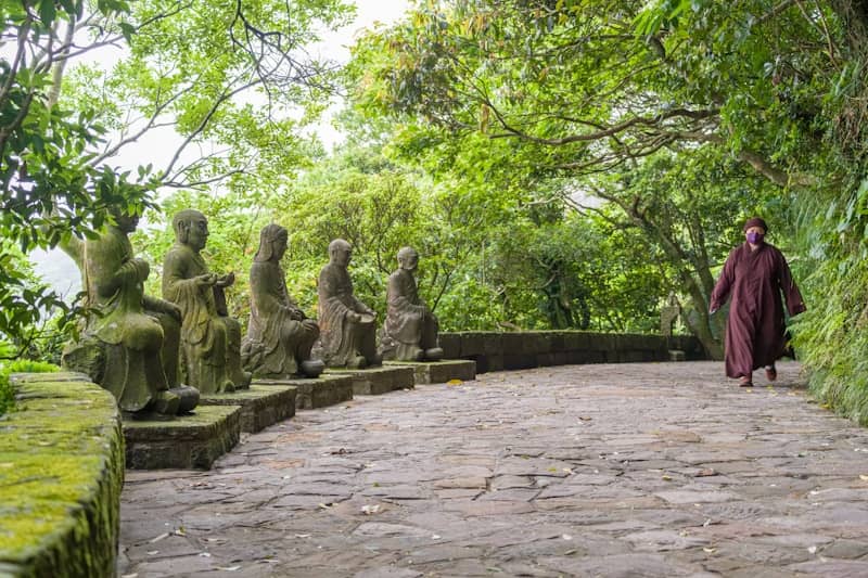 Monk walks past stone statues on a forest path.