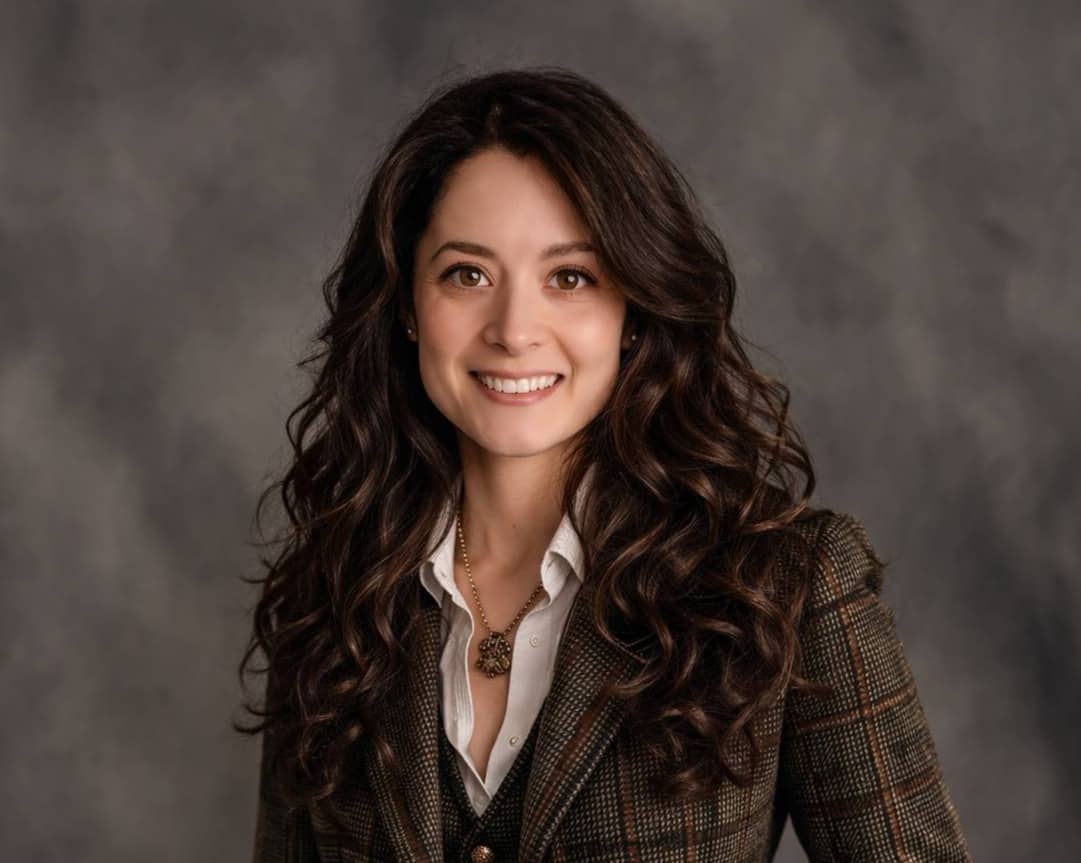 Professional portrait of a smiling woman with long dark curly hair, wearing a brown plaid blazer over a white collared shirt and dark vest, standing with one hand in her pocket against a gray studio backdrop.