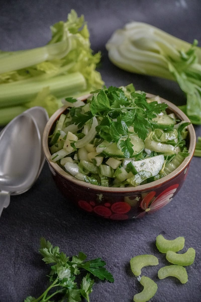 Photograph of a small floral decorated bowl containing a cucumber salad garnished with leafy green parsley. BokChoy and celery are laying on the table in the background, somewhat out of focus. a silver spoon is lying on the table next to the bowl of cucumber salad