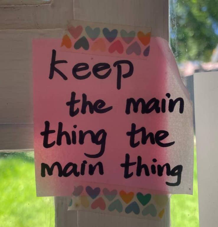 A pink sticky note taped to a windowsill with heary washi tape that reads "Keep the main thing the main thing" written with a black sharpie.