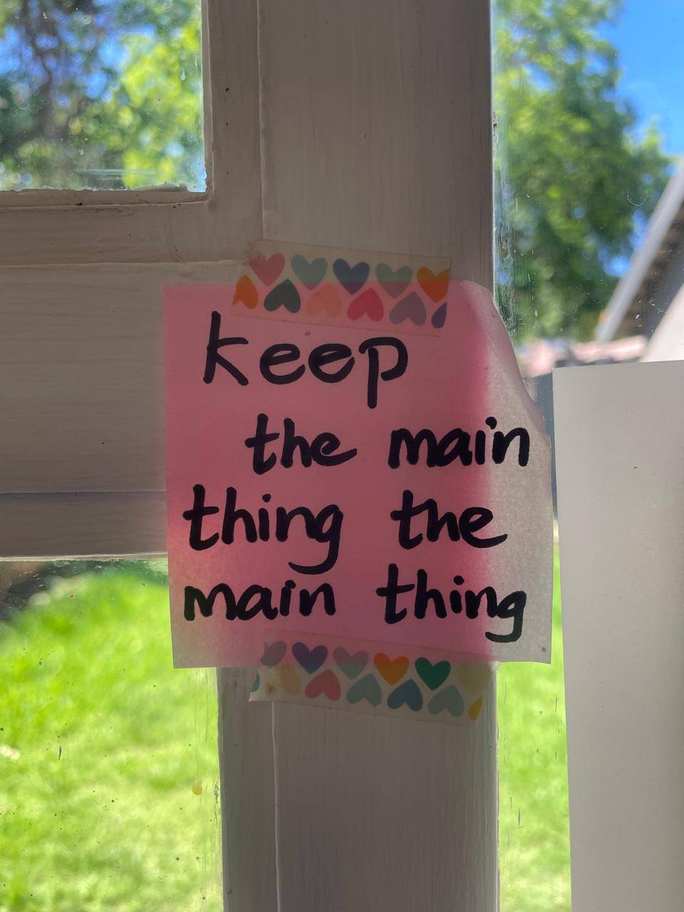 A pink sticky taped to a window with Heart patterned washi tape that reads "Keep the main thing the main thing" written in black sharpie