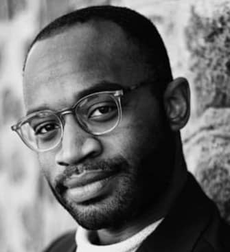 A man wearing glasses leans against a stone wall.