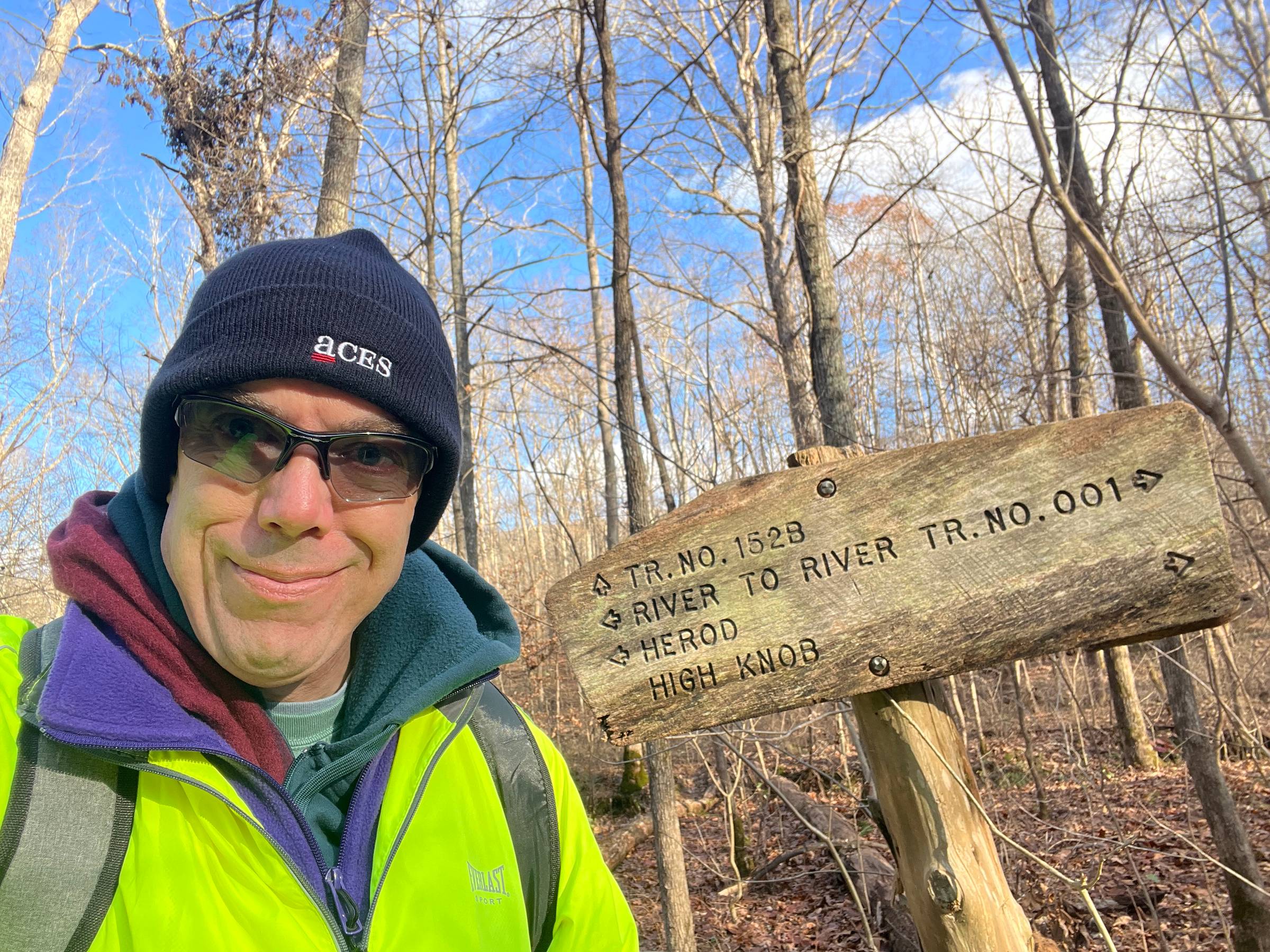 Neil takes a selfie in a leafless forest in November in front of a wooden sign noting hiking routes.