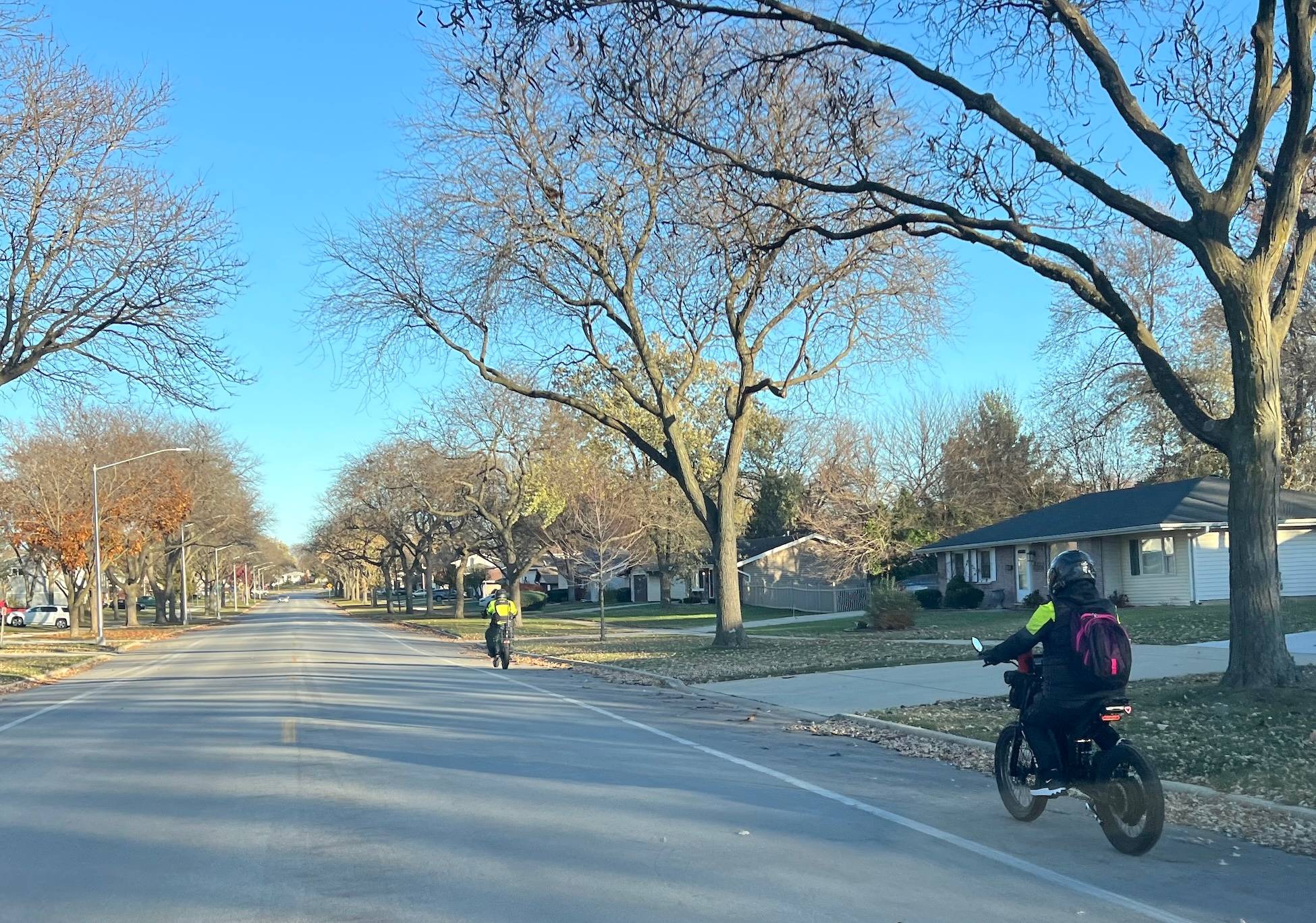 Two youths wearing motorcycle-like helmets, jackets and backpacks ride e-bikes in a bike lane along a suburban street.