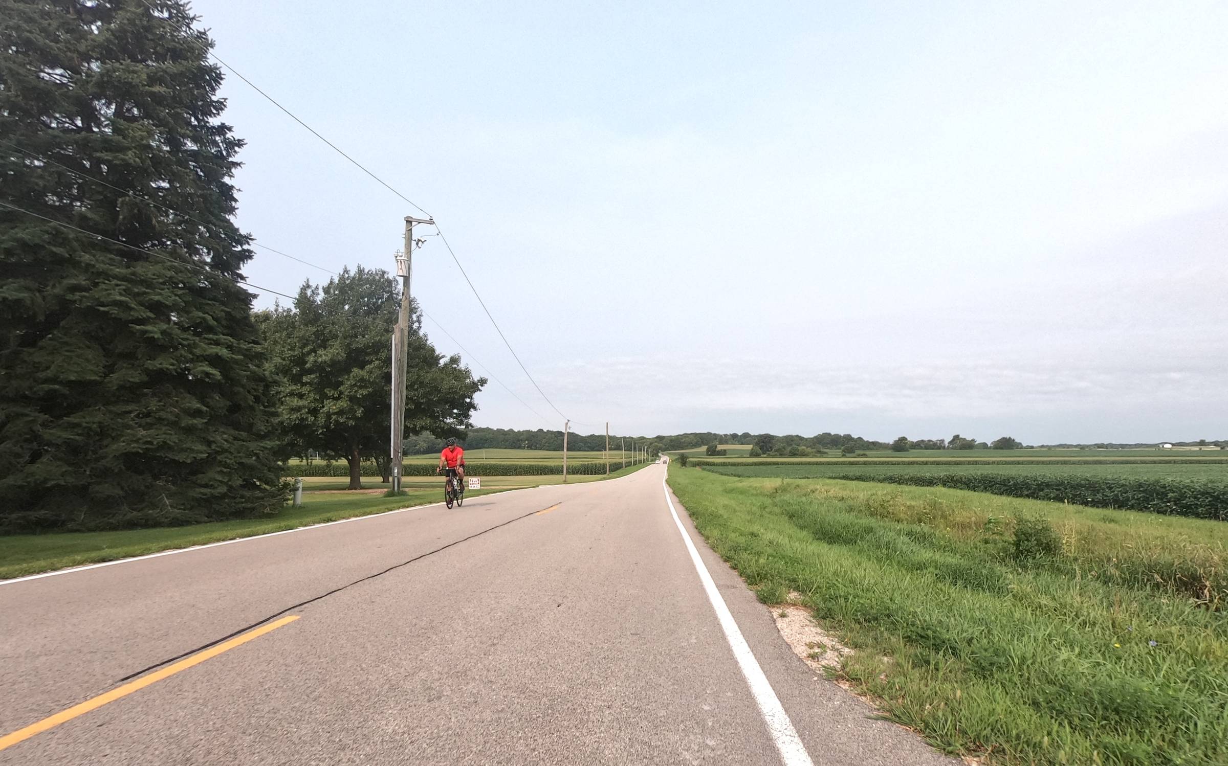 Cyclist riding on a rural road with open fields and trees.