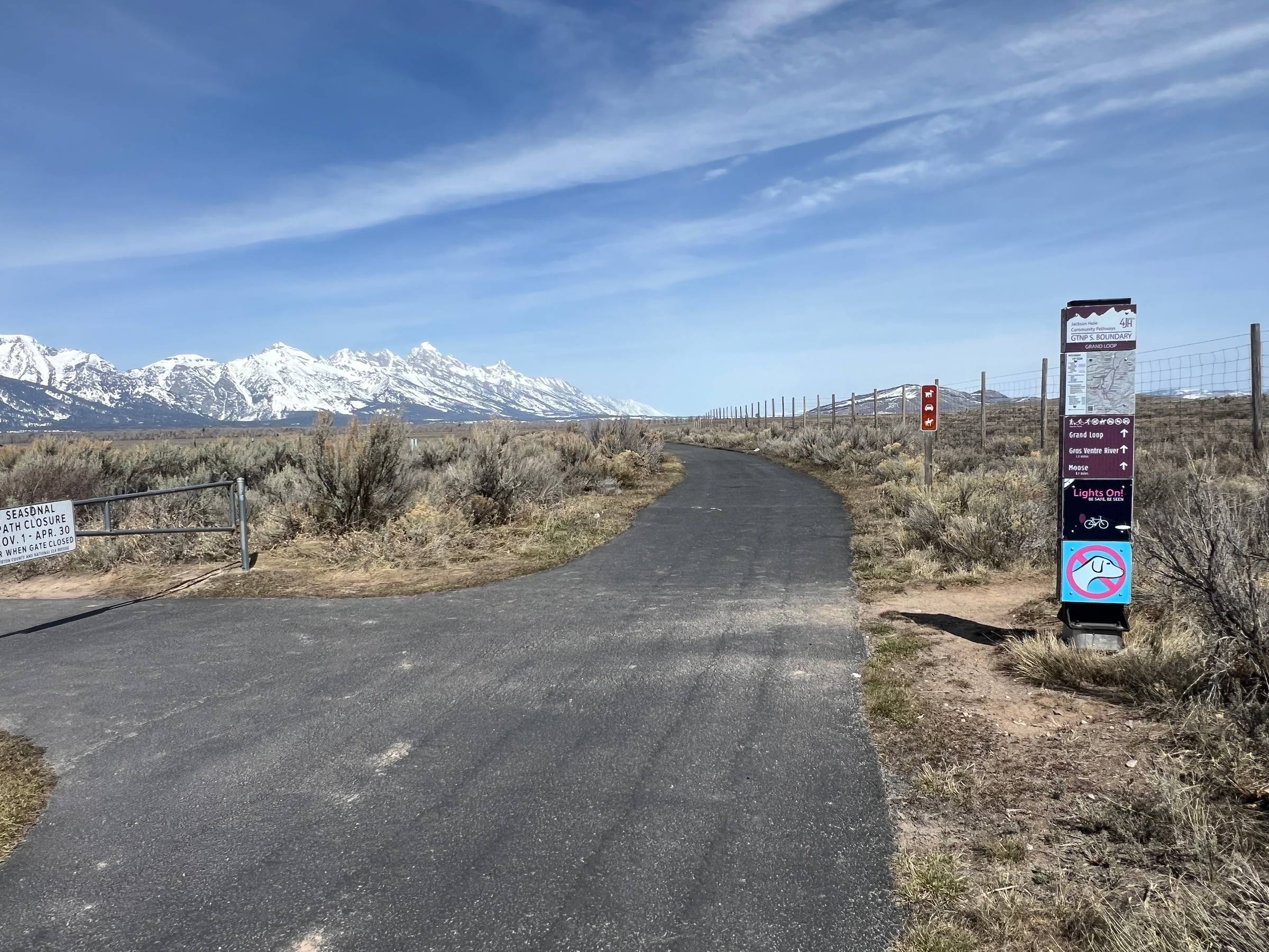 Looking down a paved trail through open prairie with trail signage up front to the right and snow-capped mountains in the left background, under blue skies.
