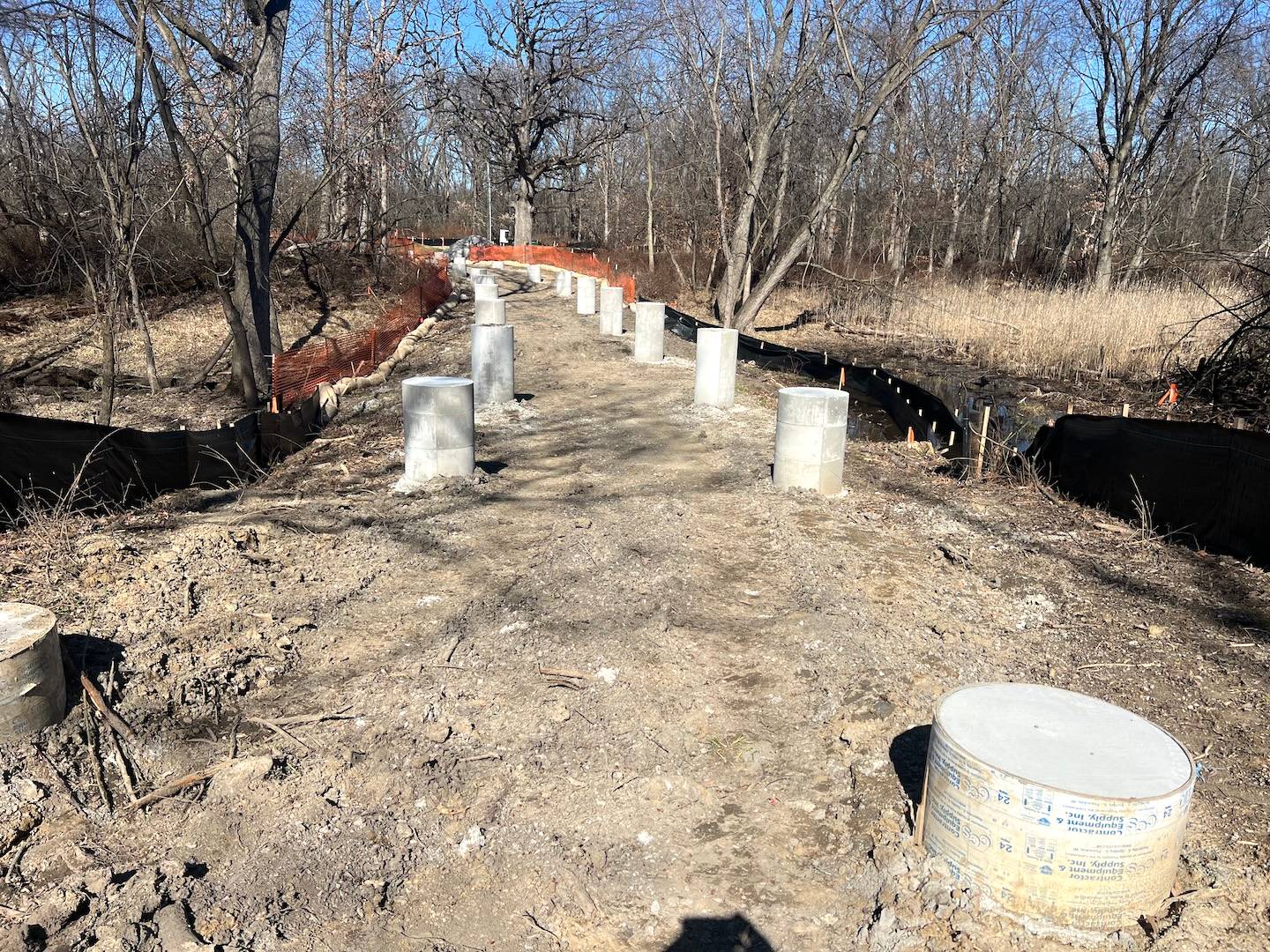 A series of short concrete support posts appear on a mud path in a forest.