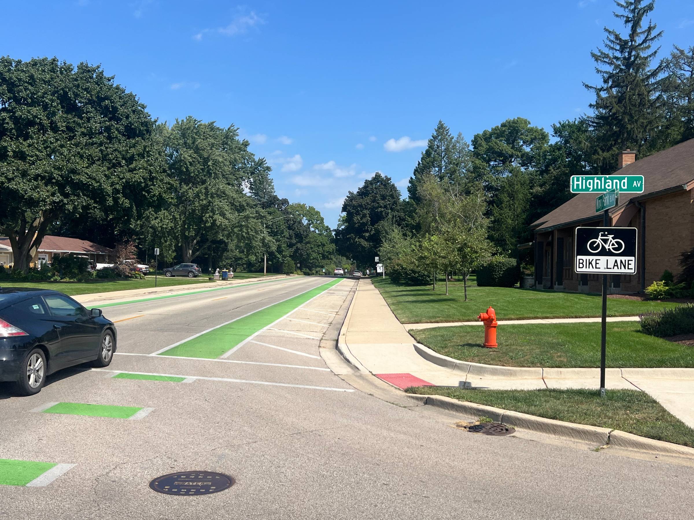 Looking down a street in a suburban neighborhood with a green painted bike lane, a Bike Route and street signs up front to the right, and trees in the background.
