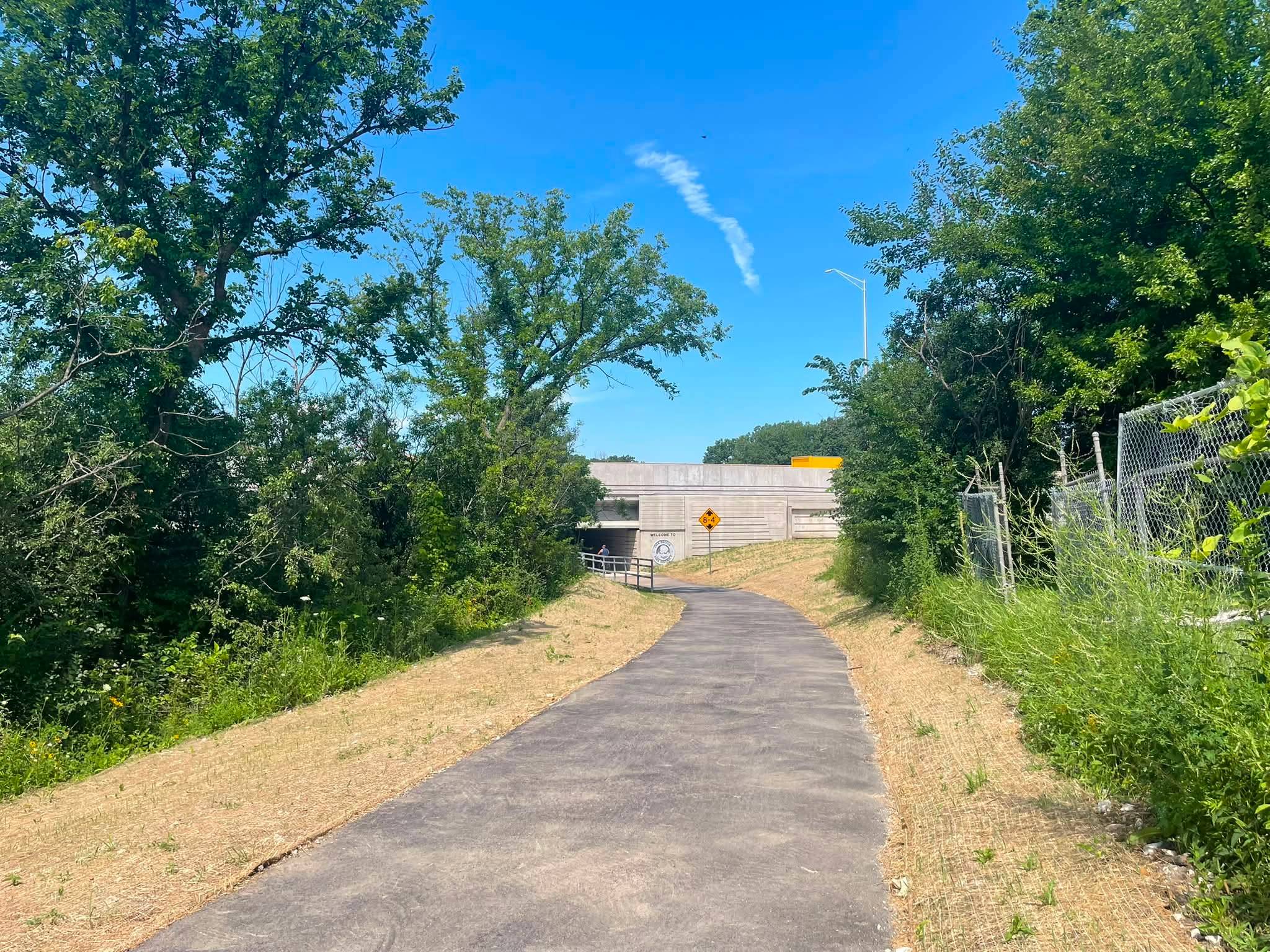 A newly paved, dustry trail curves through new lawn and rough bushes toward an underpass under a blue sky