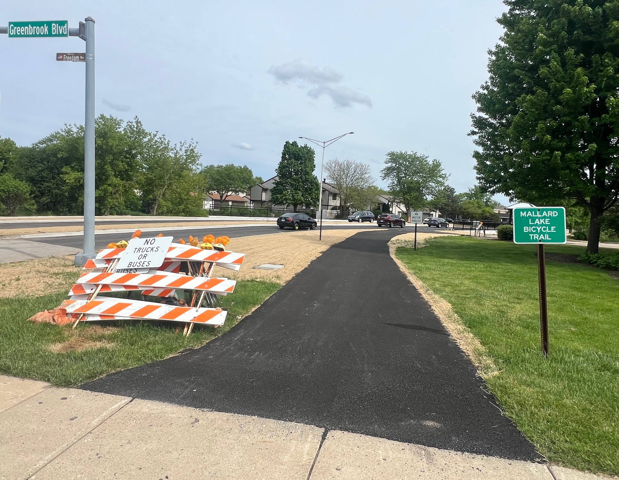 A bike trail with new, black asphalt runs through a suburban neighborhood with removed construction signs off to the side and a Mallard Lake Bicycle Trail sign.
