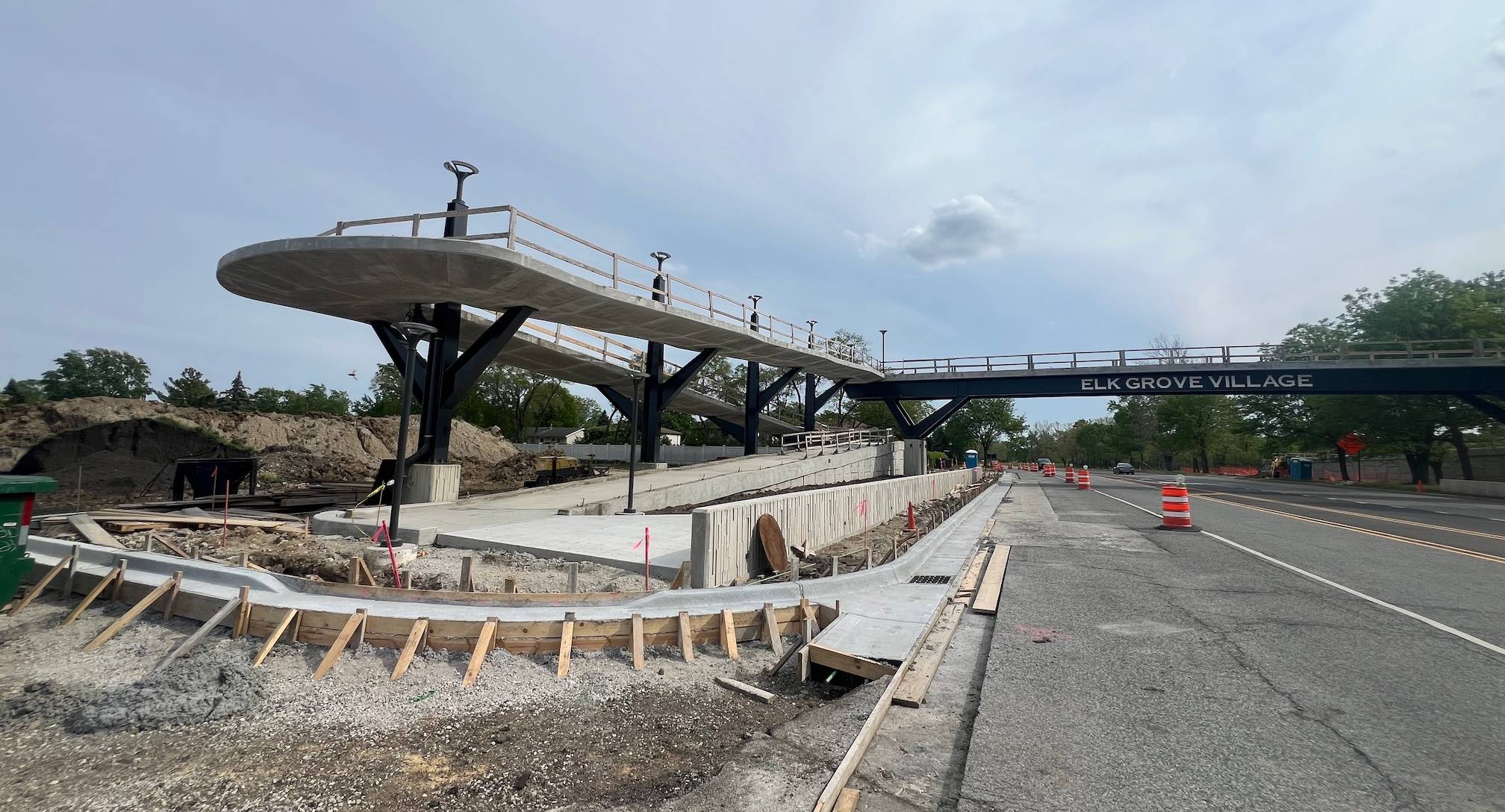 A winding ramp up to a bike-pedestrian bridge labeled Elk Grove Village is under construction by a suburban road