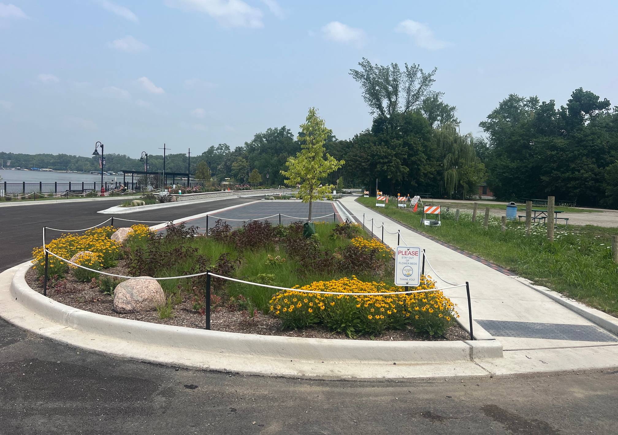 A sidewalk runs on the right side past a garden in a median, ahead of a newly paved road with another median in the background as well as a lake.