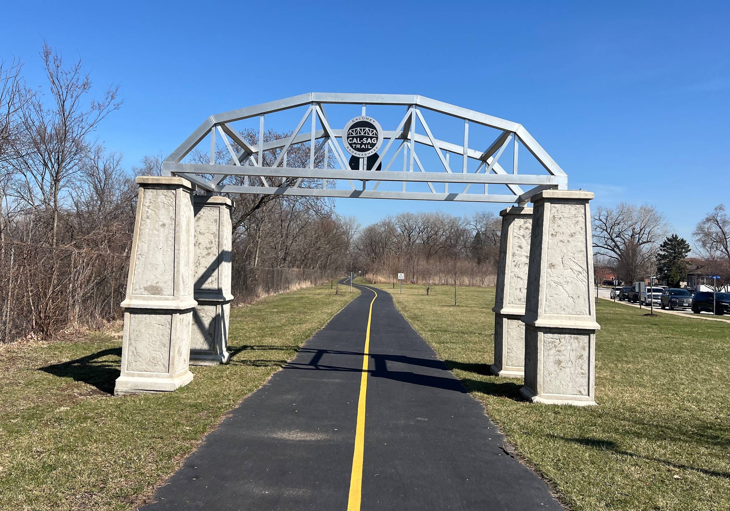 A gateway made to look like a small bridge appears over a newly paved, striped asphalt trail through a park in a suburb