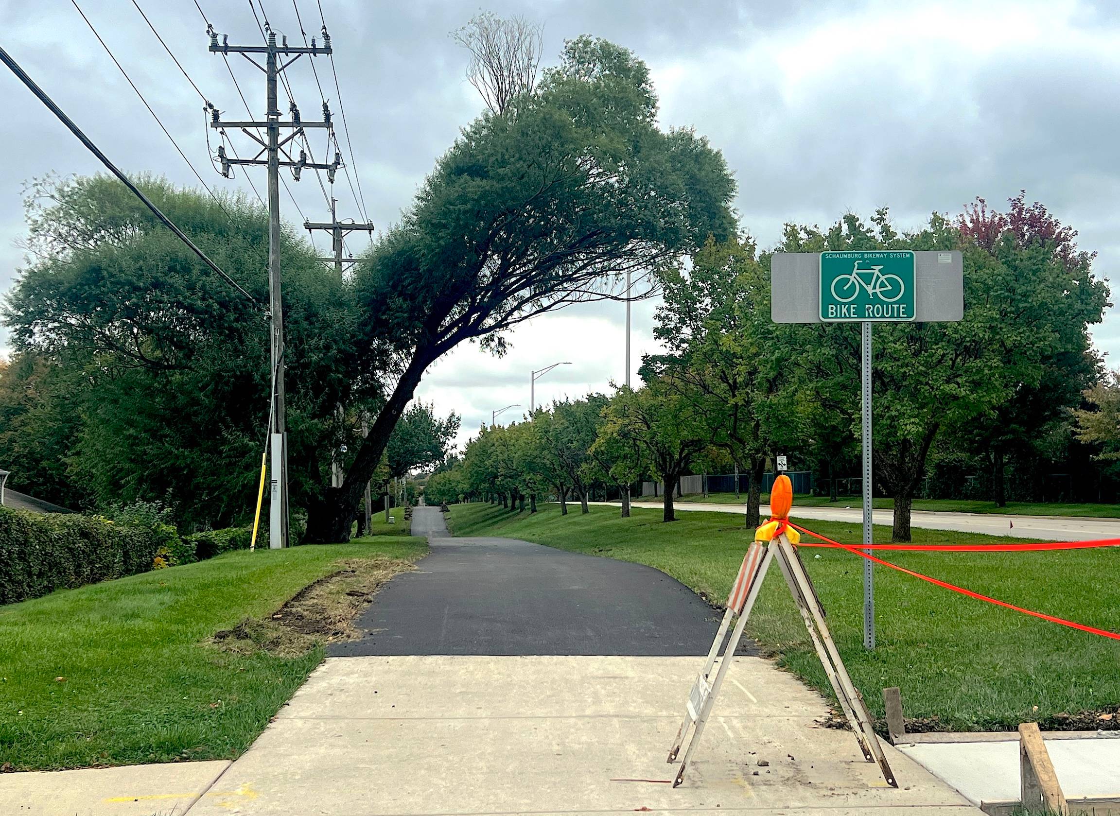 A bike path through a green suburban neighborhood has new black asphalt behind a construction horse and Bike Route sign